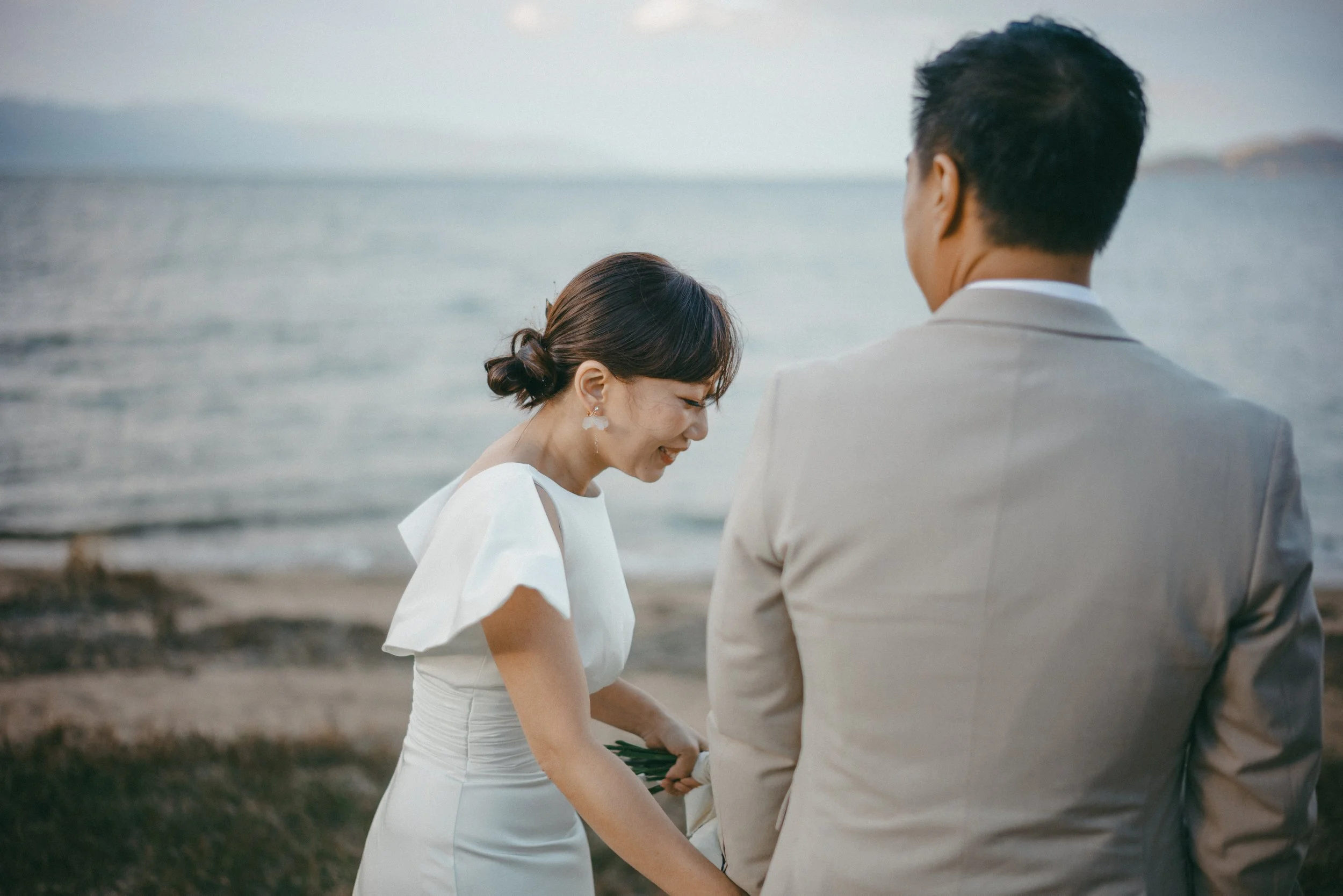 A woman in a white dress and a man in a light suit stand on the beach by the water, holding hands and smiling, during sunset.