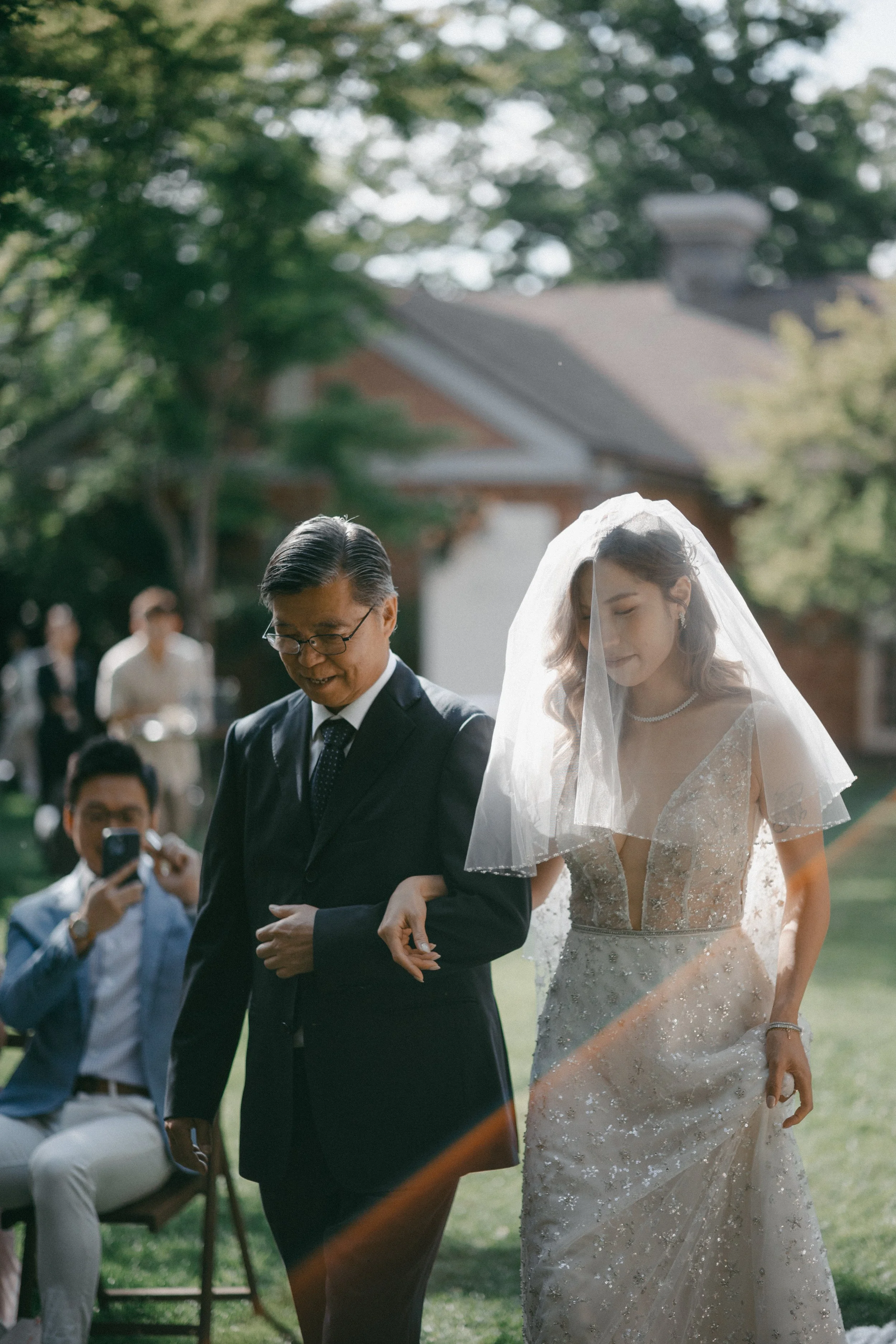 Bride walking down the aisle with her father during an outdoor wedding ceremony on a sunny day.