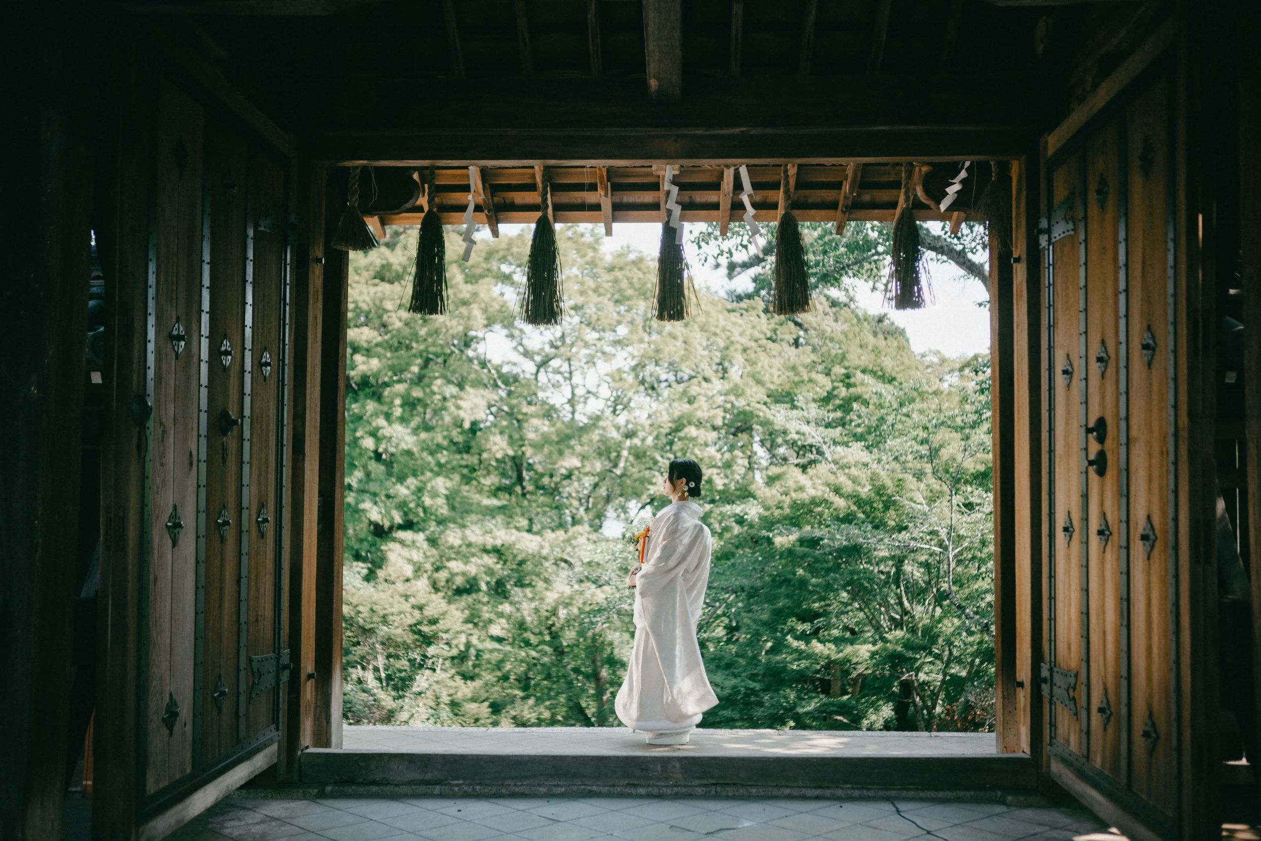 A woman in traditional white Japanese wedding kimono standing on a wooden porch, holding a small bouquet, looking towards lush green trees outside.