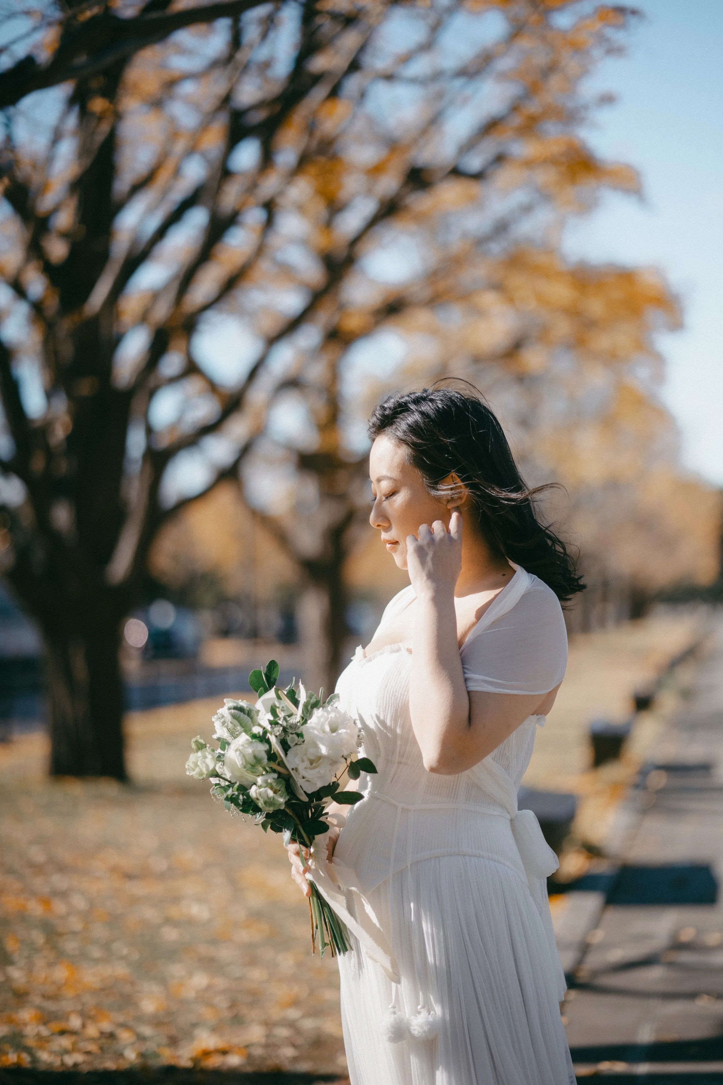 A woman in a white dress holding a bouquet of white flowers, standing outdoors with autumn trees in the background.