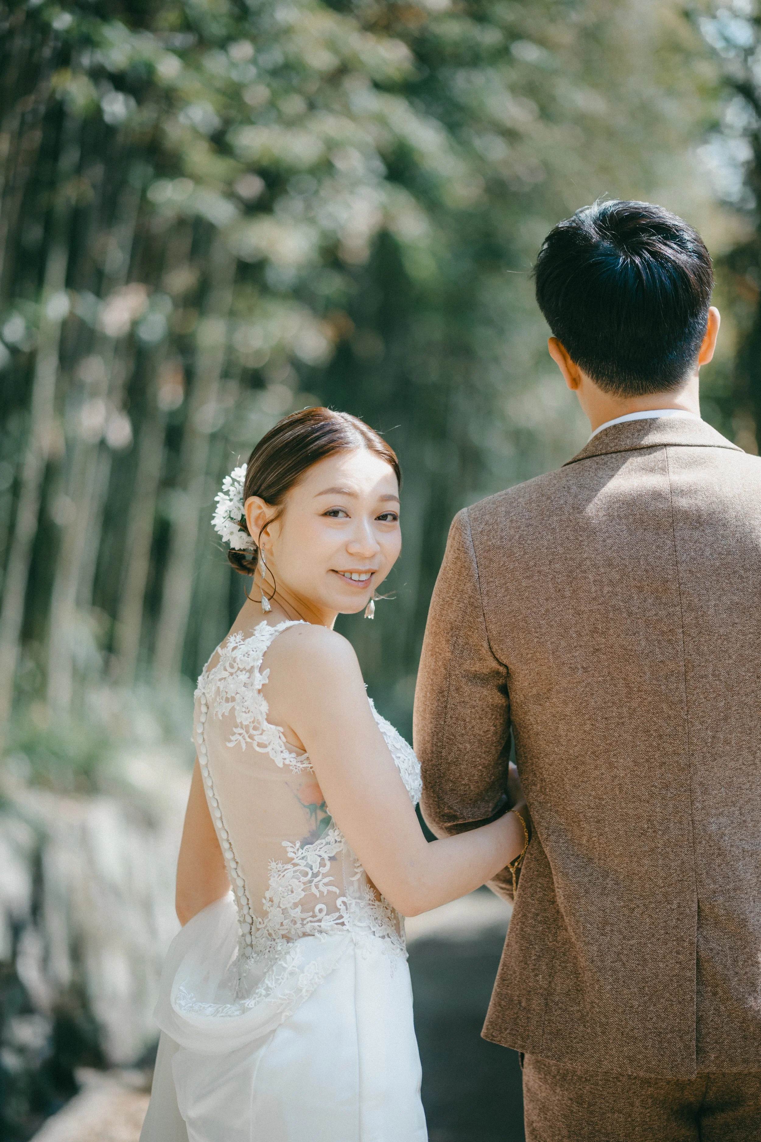 A smiling bride with a flower in her hair holding hands with a groom, taken outdoors with trees in the background.