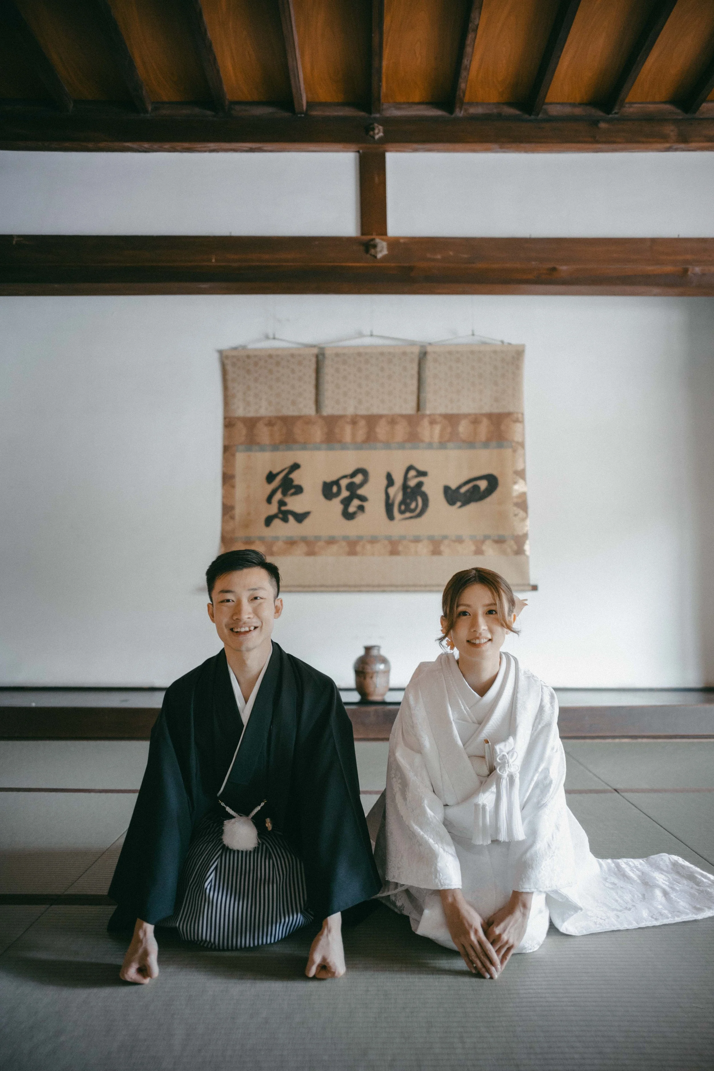 A Japanese man and woman dressed in traditional wedding attire, kneeling on tatami mats in a traditional room, with a scroll and pottery in the background.