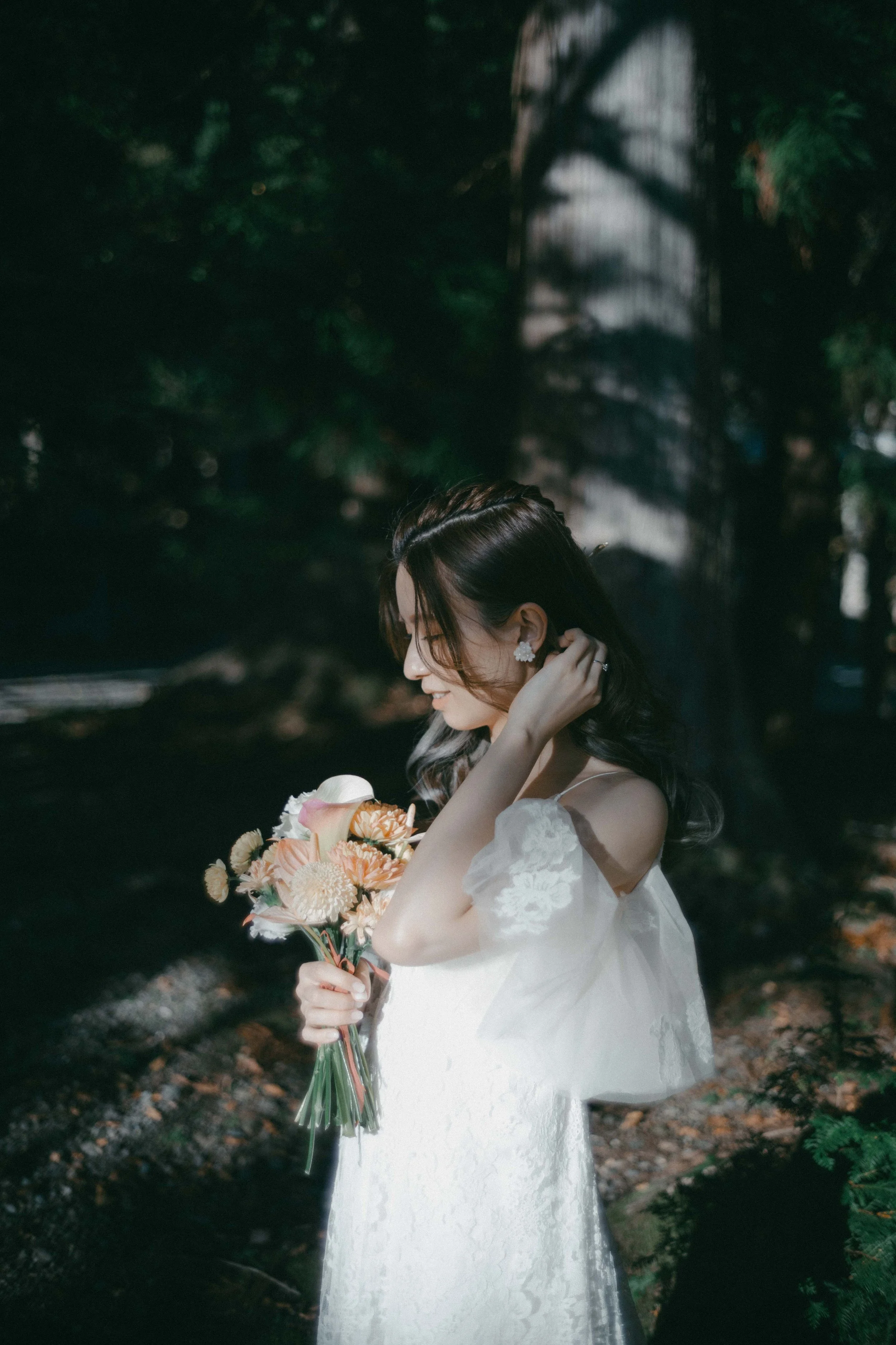 A woman in a white wedding dress holding a bouquet of pink and white flowers, standing outdoors near a tree with sunlight casting shadows.
