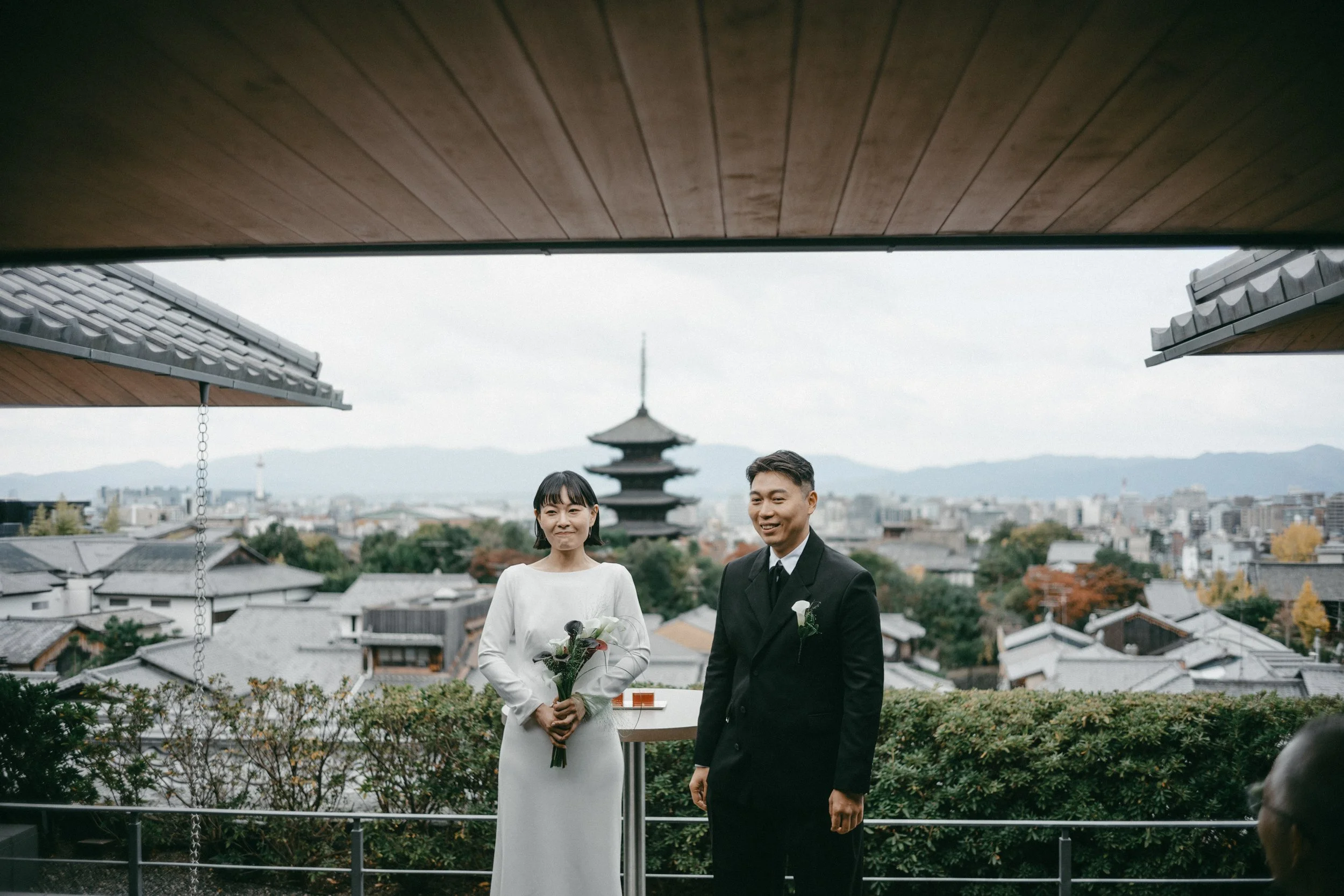 A couple getting married outdoors on a balcony with a view of a traditional Japanese pagoda and city skyline in the background, the bride in a white dress holding a bouquet, and the groom in a black suit.