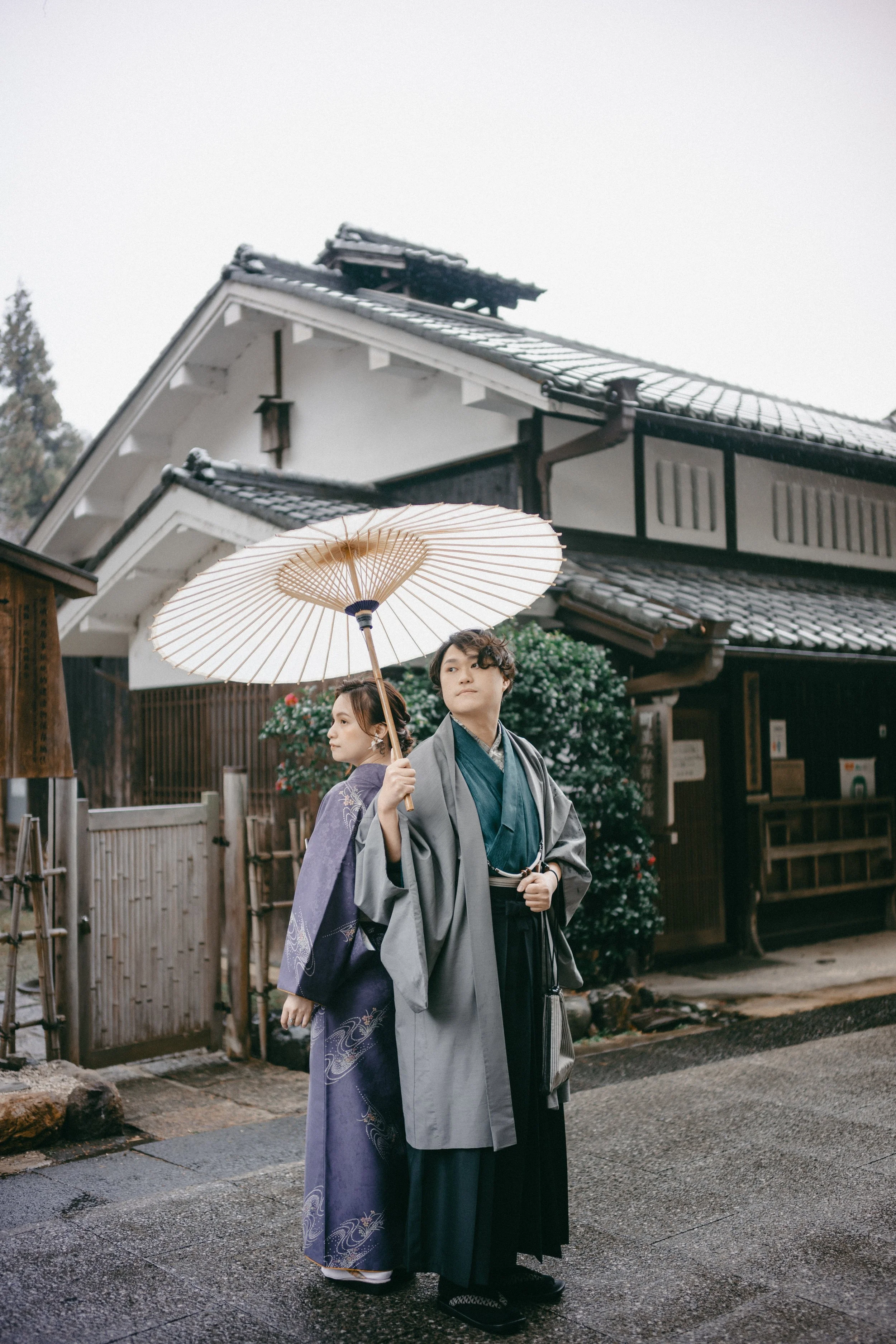 Two people dressed in traditional Japanese clothing stand outside a historic building, one holding a paper parasol.