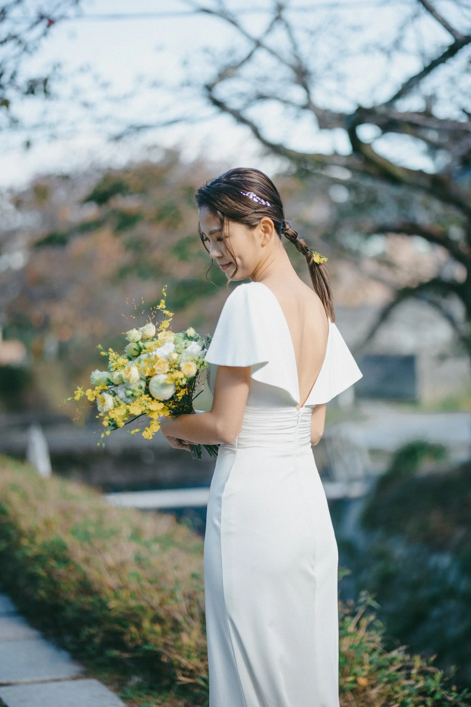 A woman in a white dress holding a bouquet of yellow and white flowers outdoors. She has dark hair styled in a braided ponytail with floral accessories, standing in front of trees with bare branches.