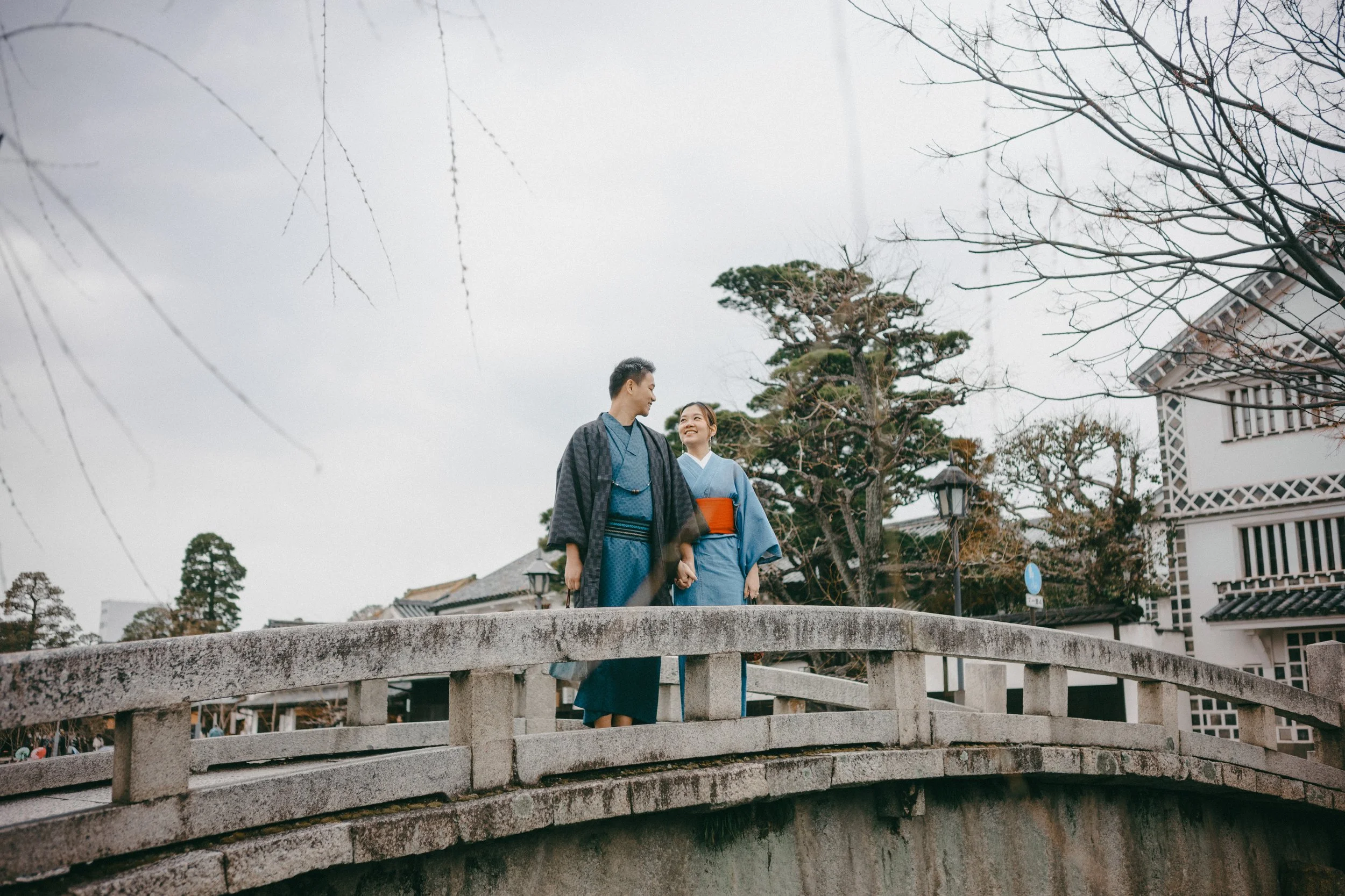 A young couple in traditional Japanese kimonos walking hand in hand across a stone bridge, smiling at each other, with trees and classic buildings in the background on a cloudy day.