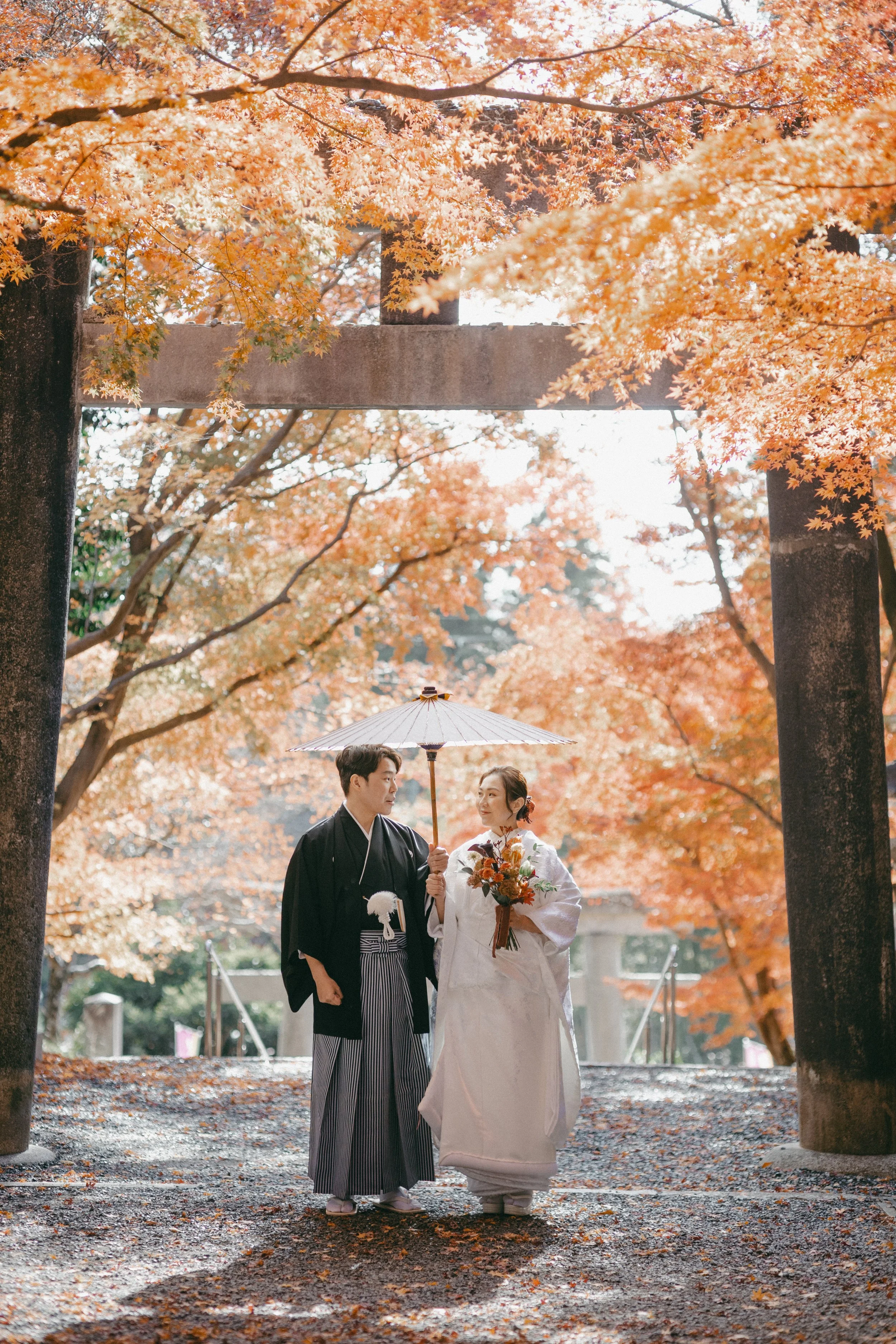 A couple dressed in traditional Japanese wedding attire, standing under an umbrella amidst vibrant autumn foliage, with a torii gate in the background.