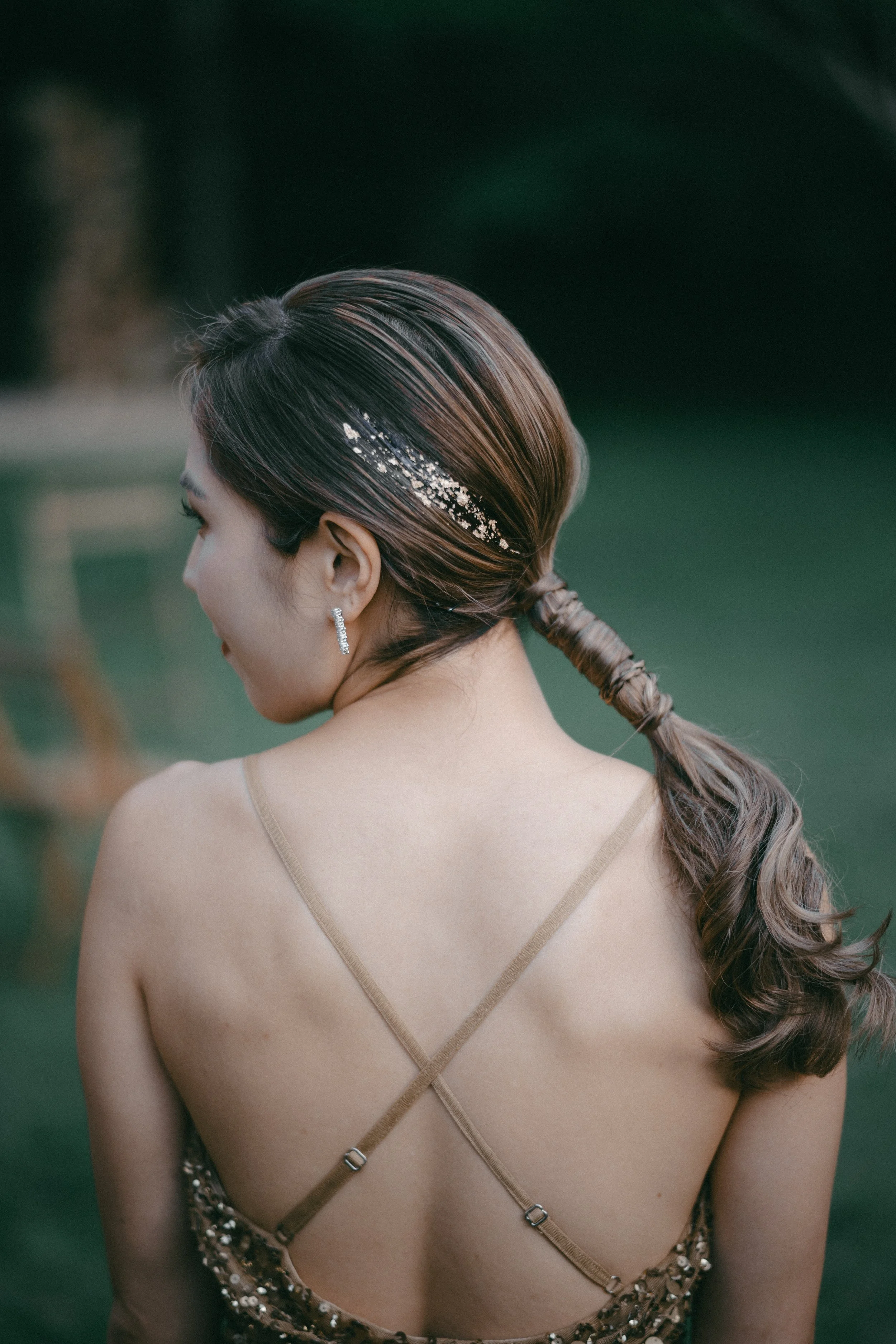 A woman with long, styled hair wearing a sparkling hair accessory, earrings, and a dress with thin straps, is photographed from behind outdoors.