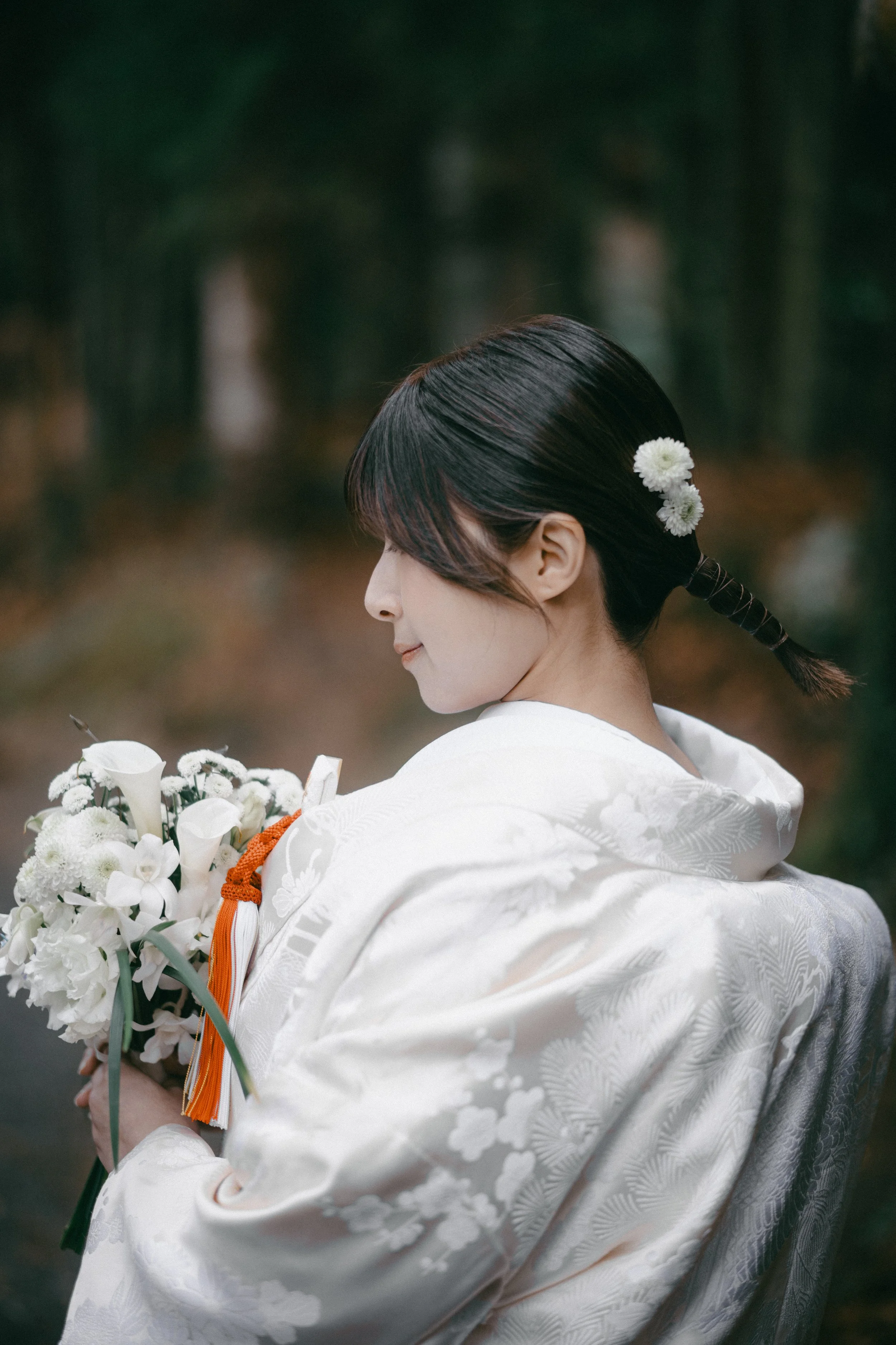 A woman in traditional white Japanese wedding attire holding a bouquet of white flowers, with her hair styled in a braided updo adorned with white flowers, standing outdoors with a blurred background of trees.
