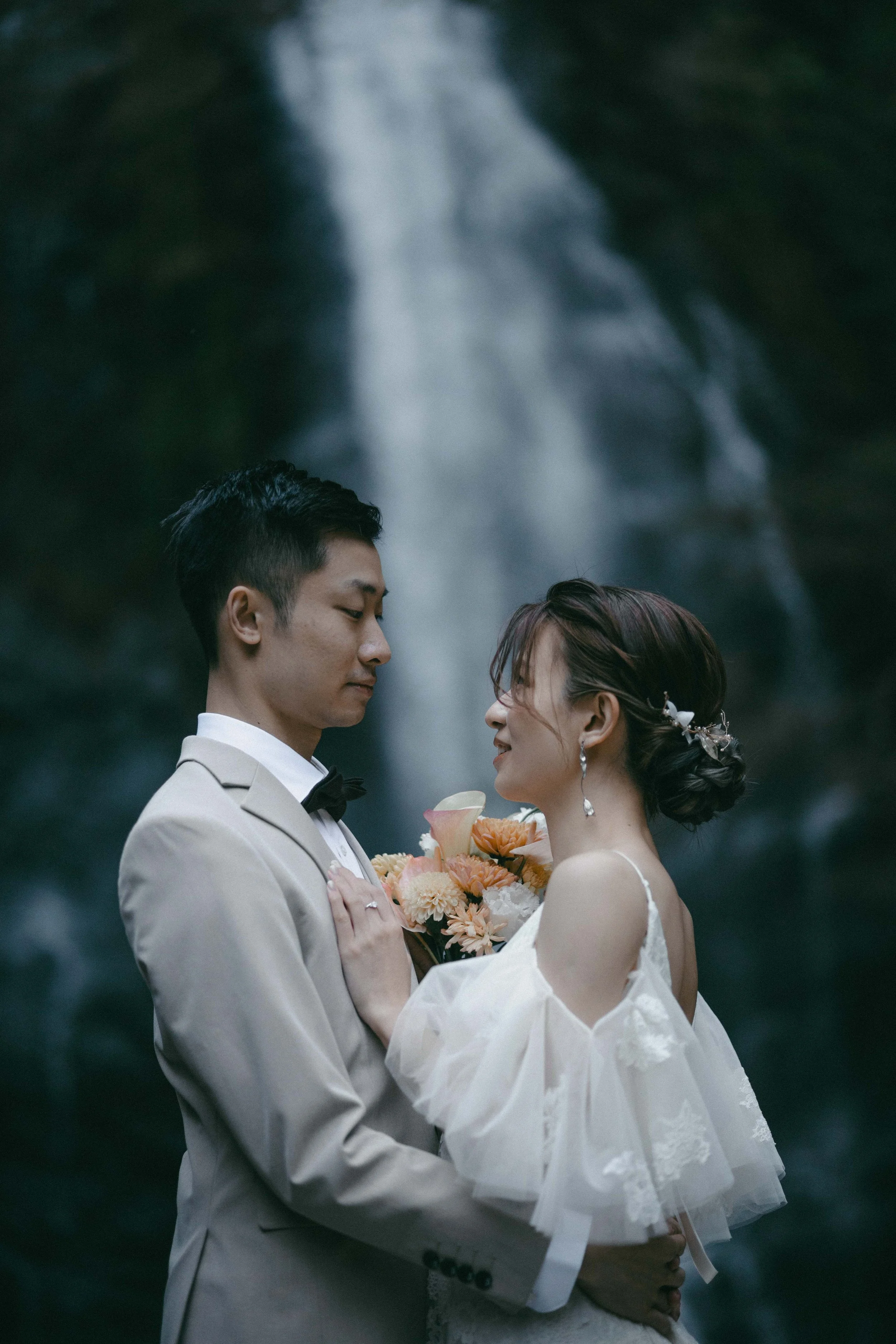 A bride and groom standing close together, looking into each other's eyes, with a waterfall in the background. The bride is holding a bouquet of flowers.