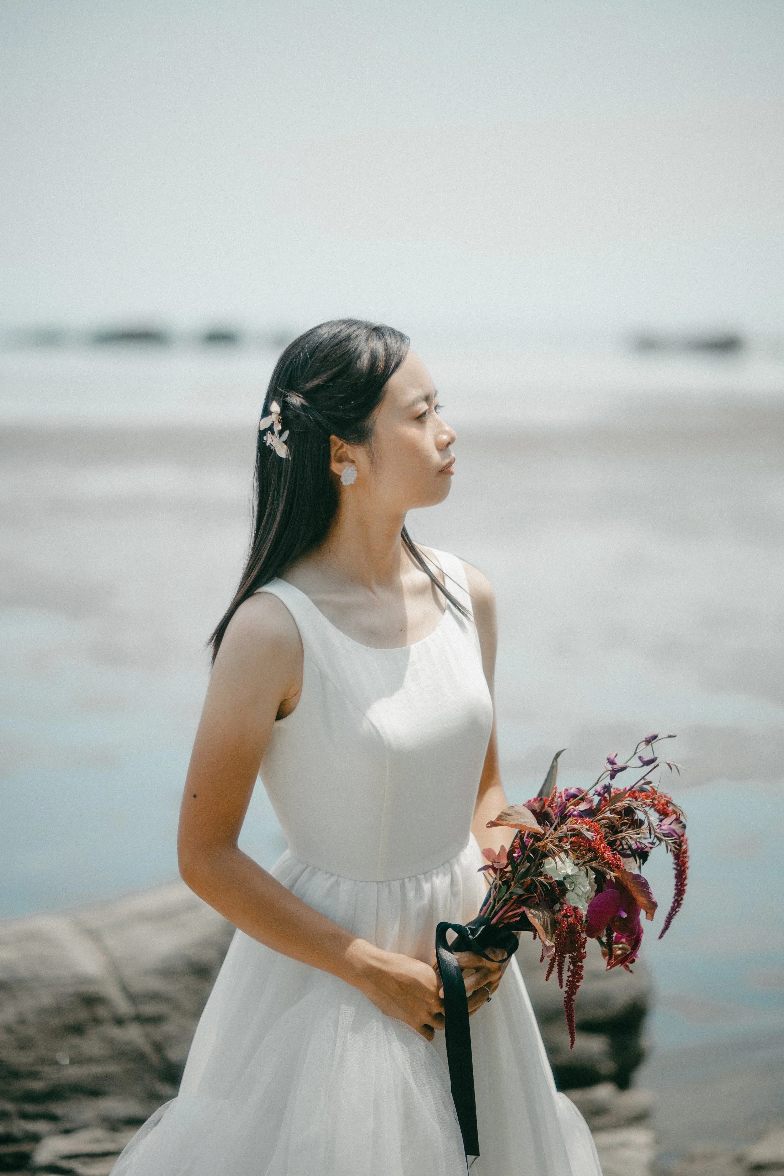 A woman in a white wedding dress holding a bouquet of flowers on a beach.