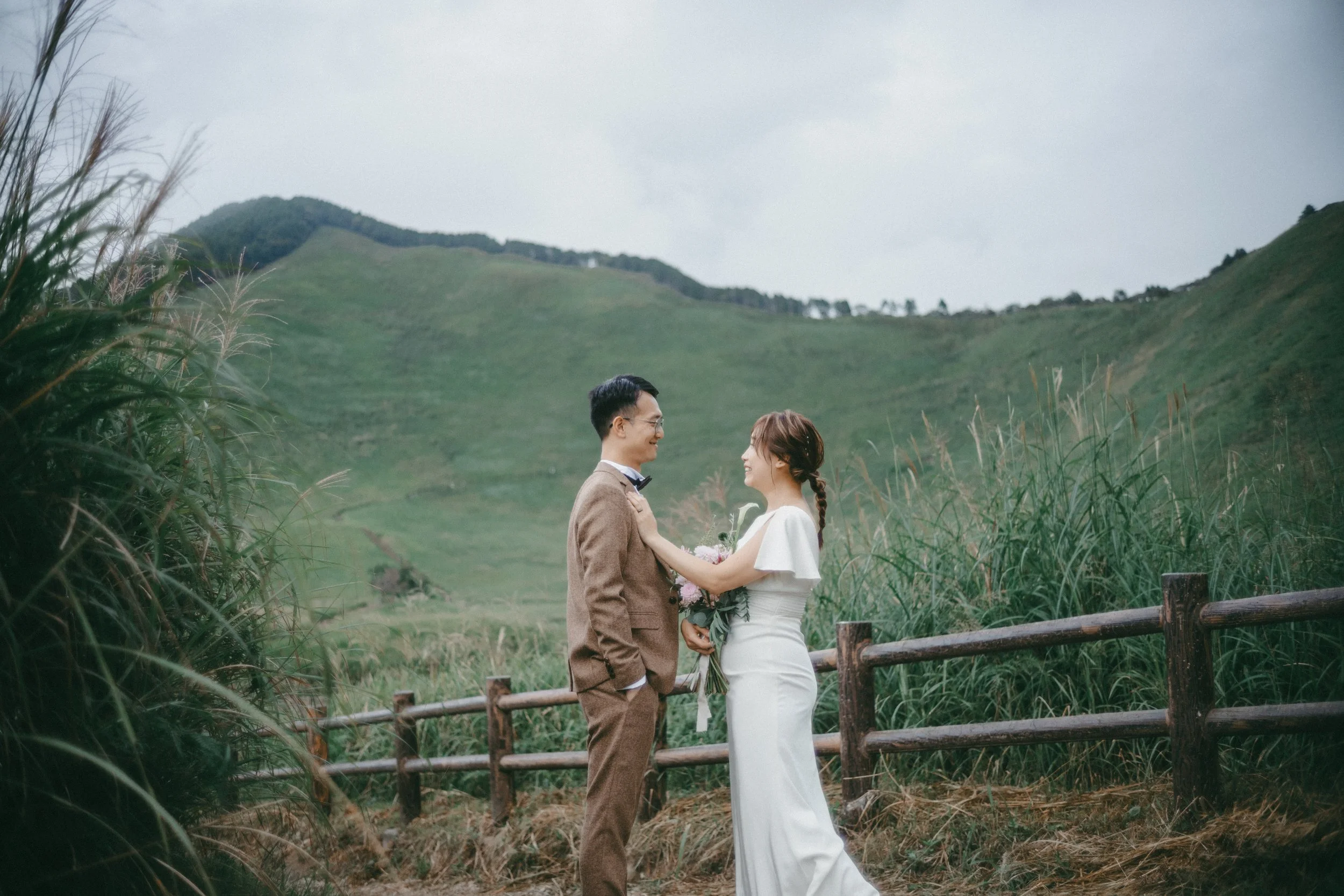 A couple in wedding attire standing on a path with a rustic wooden fence, surrounded by tall grass and green hills in the background, smiling at each other.