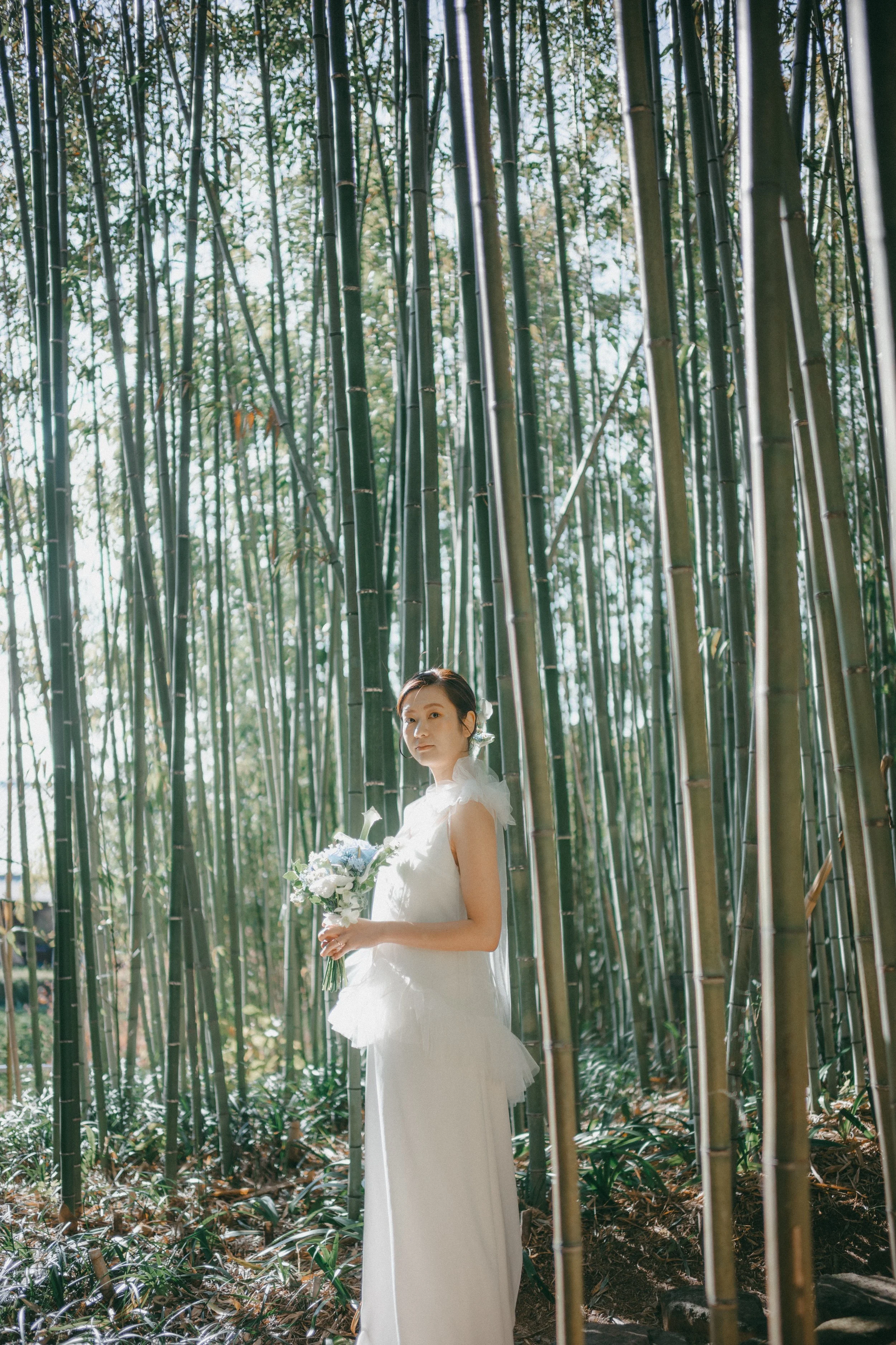 A woman in a white wedding dress standing in a bamboo forest holding a bouquet of white and blue flowers.