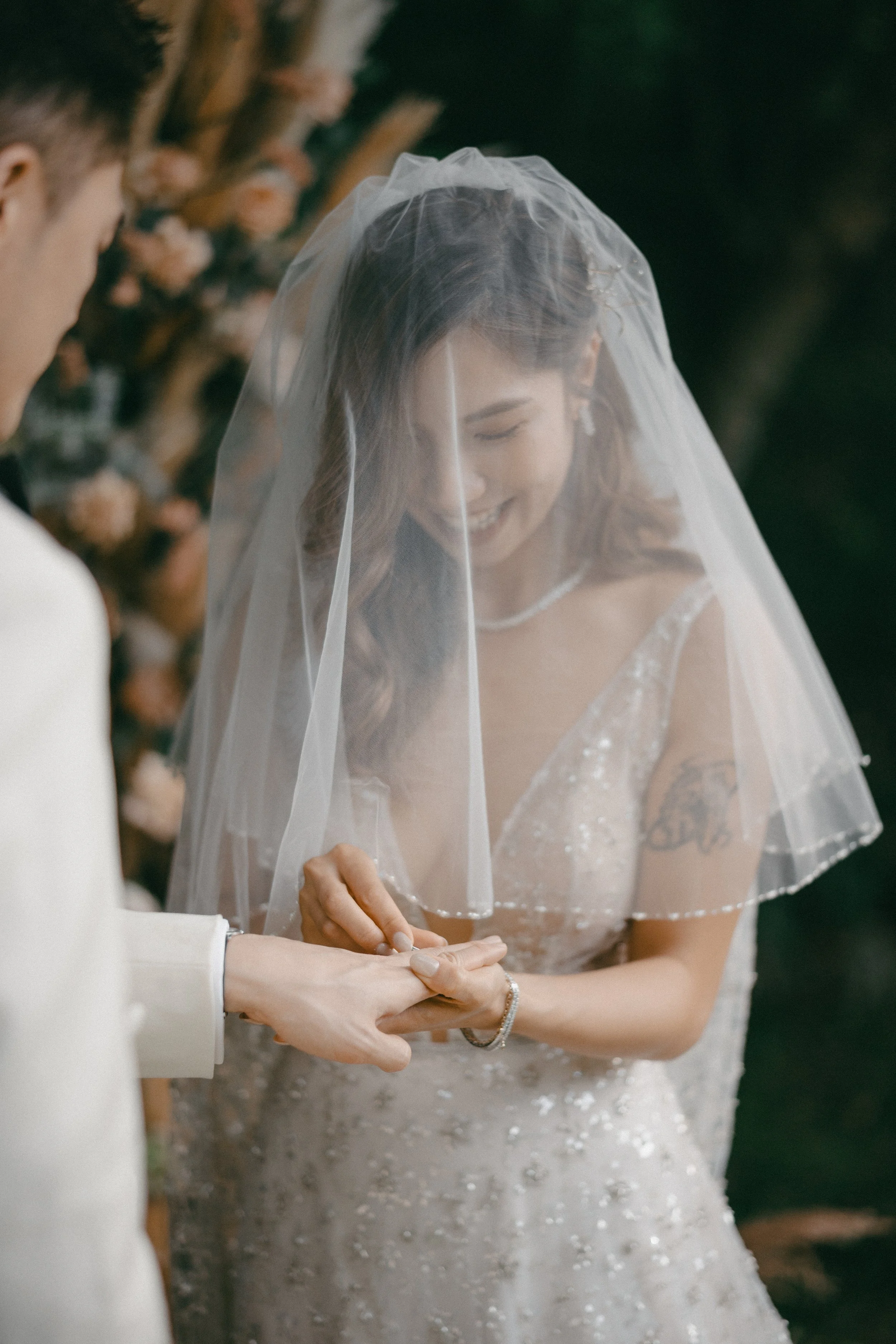 A bride and groom exchanging wedding rings outside, with the bride smiling and wearing a veil and wedding dress.