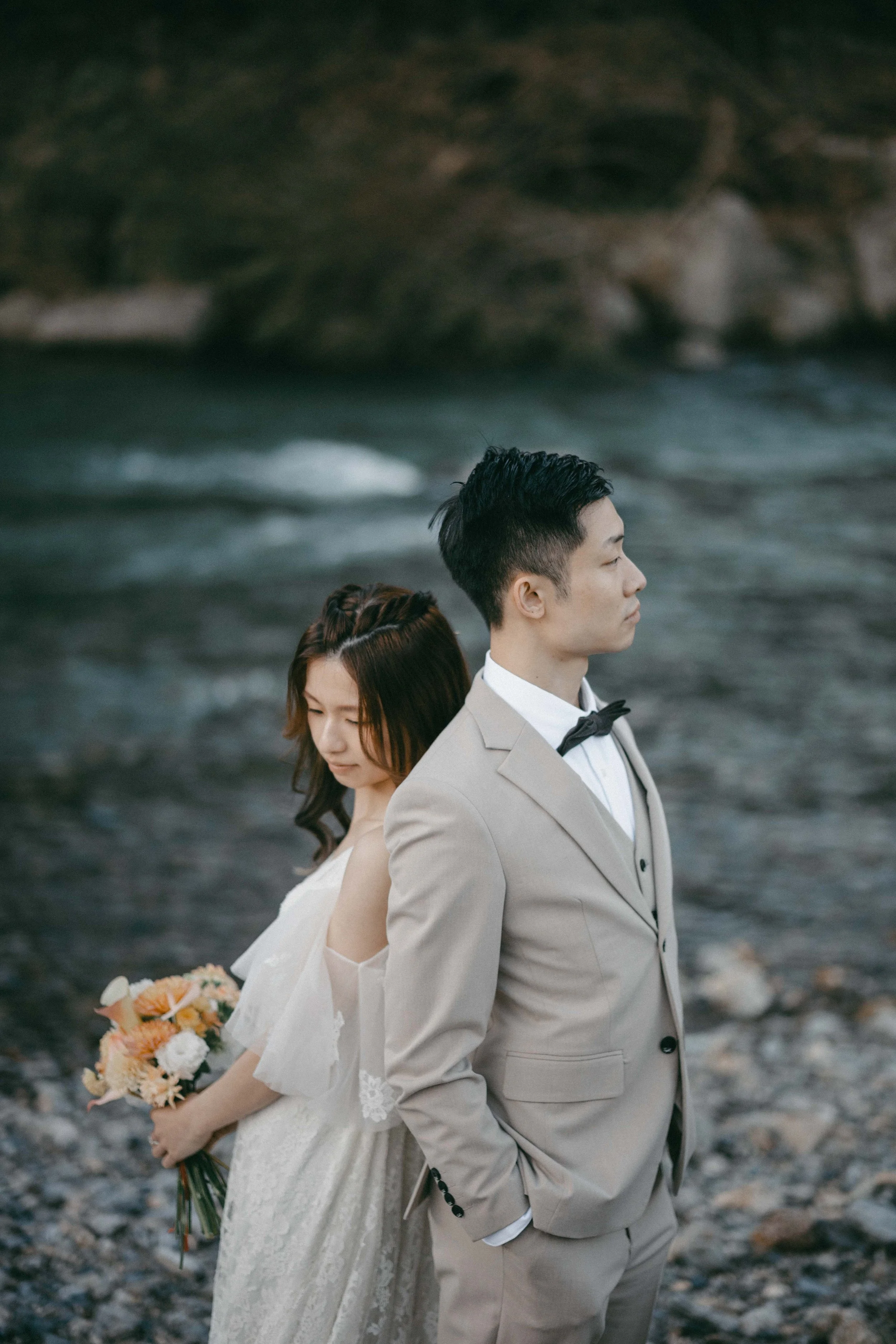 A bride and groom standing back to back on a rocky beach, with the bride holding a bouquet of flowers and looking downward, and the groom looking to the side with hands in his pockets.