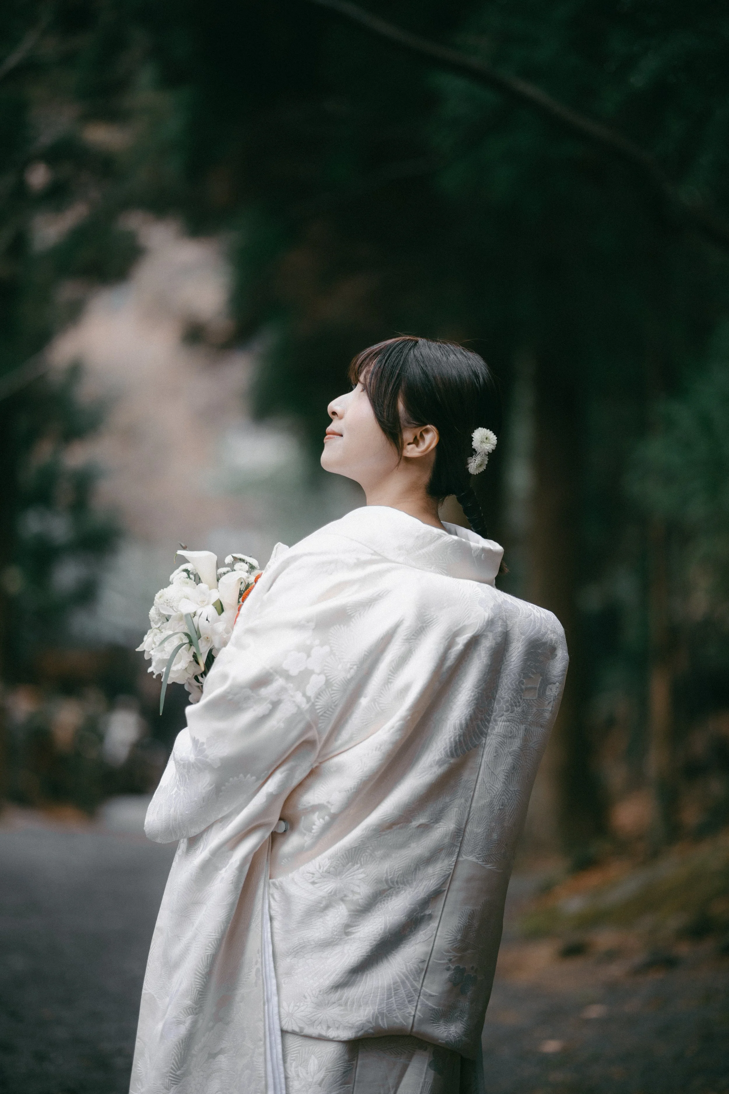 A woman dressed in a traditional white kimono, holding a bouquet of white flowers, standing outdoors with a serene expression in a wooded area.