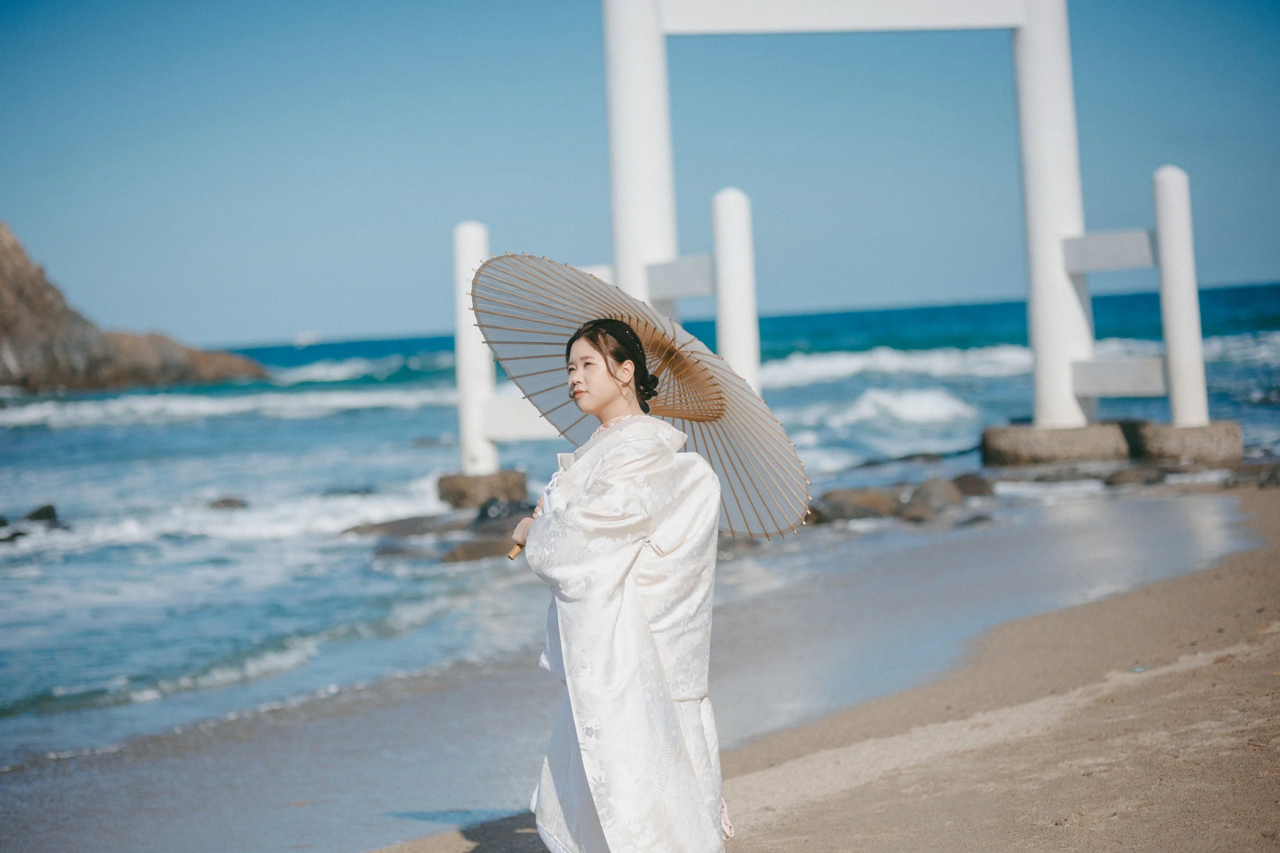 A woman in traditional white clothing holding a parasol on a beach with waves and a distant rock formation, and a white torii gate in the background.