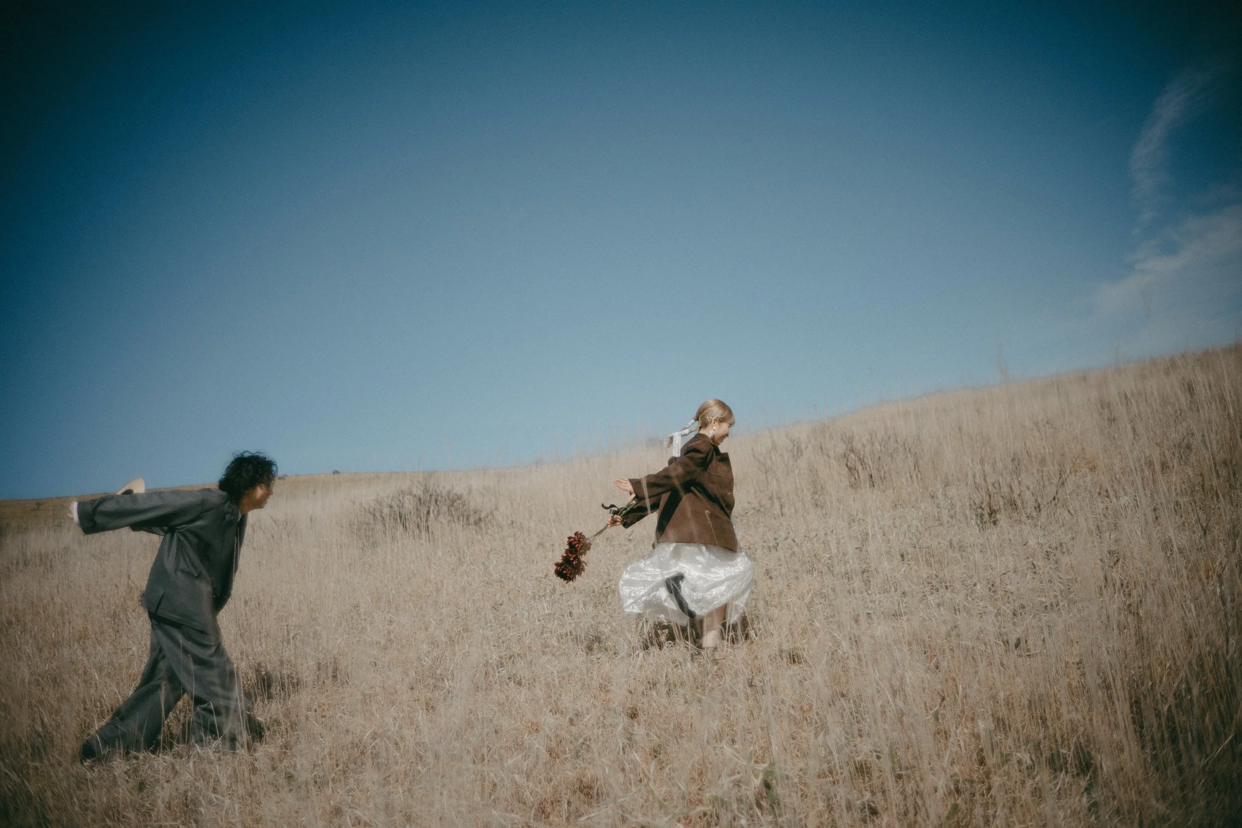 A woman in a white dress and brown jacket holding a bouquet of flowers is walking up a dry grassy hill, with a man in a dark suit following behind her under a blue sky.