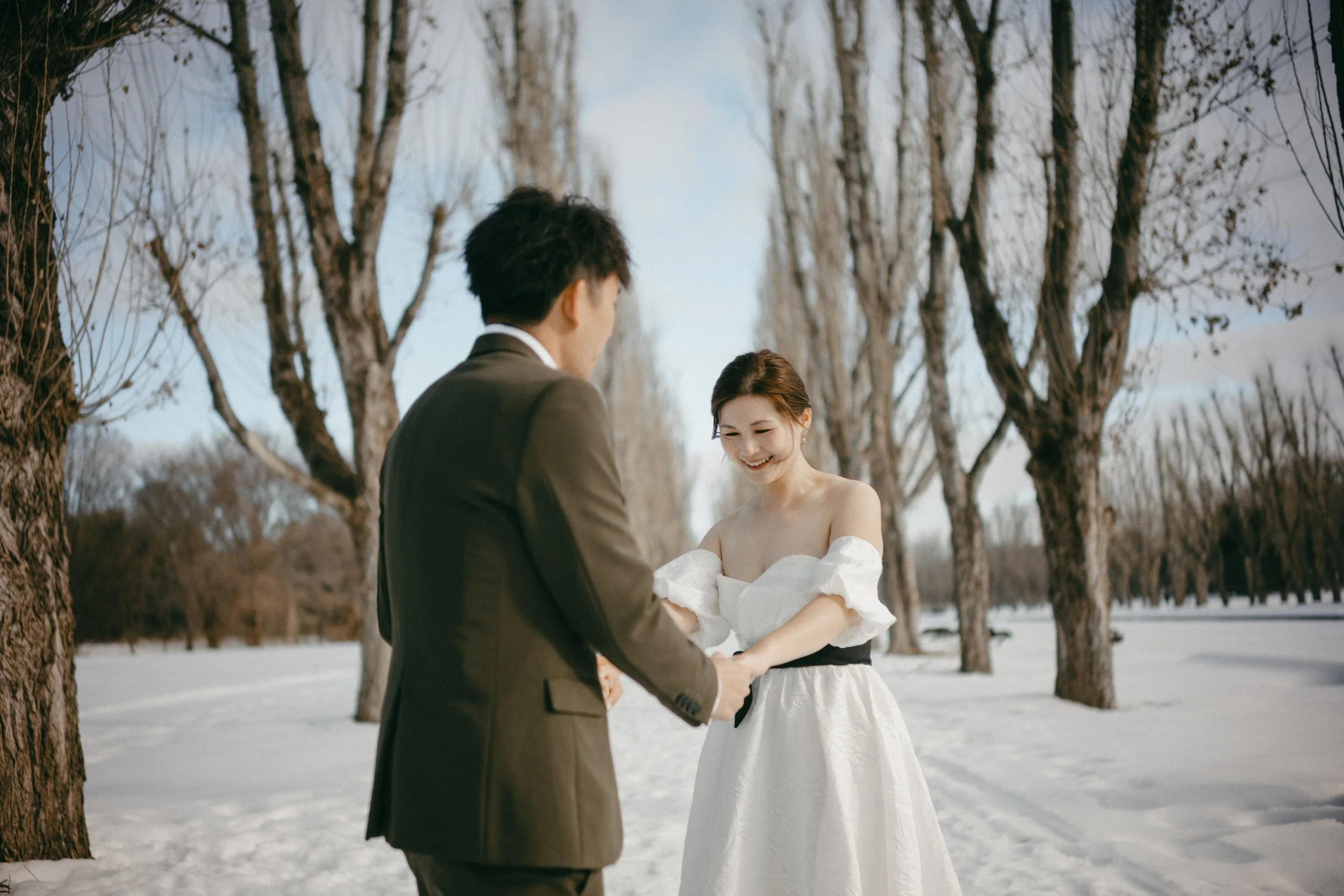 A couple dressed in wedding attire exchanging rings in a snowy outdoor setting with bare trees.