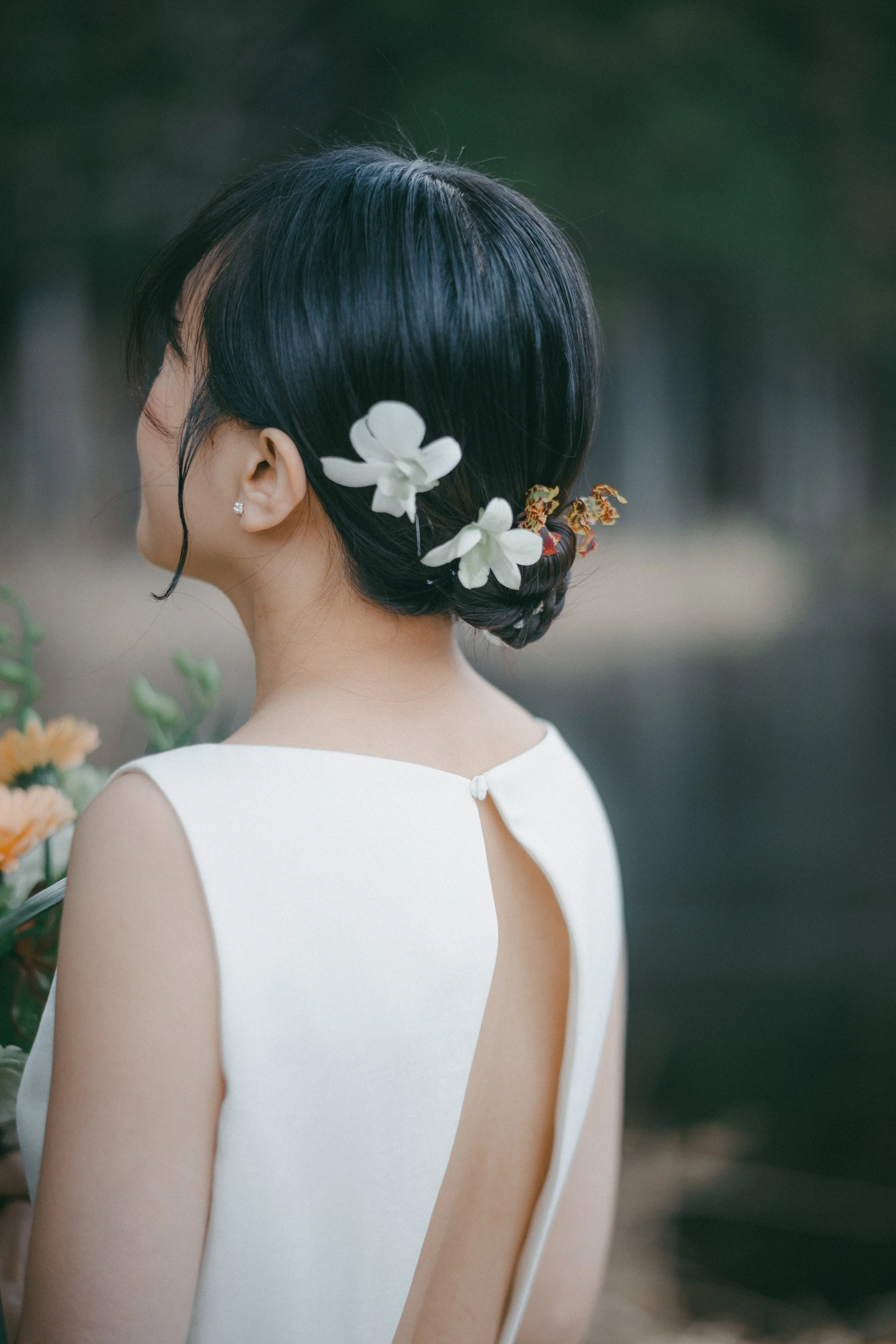 A woman with black hair styled in an elegant updo decorated with white flowers, wearing a white dress with a keyhole back, standing outdoors with a blurred natural background.