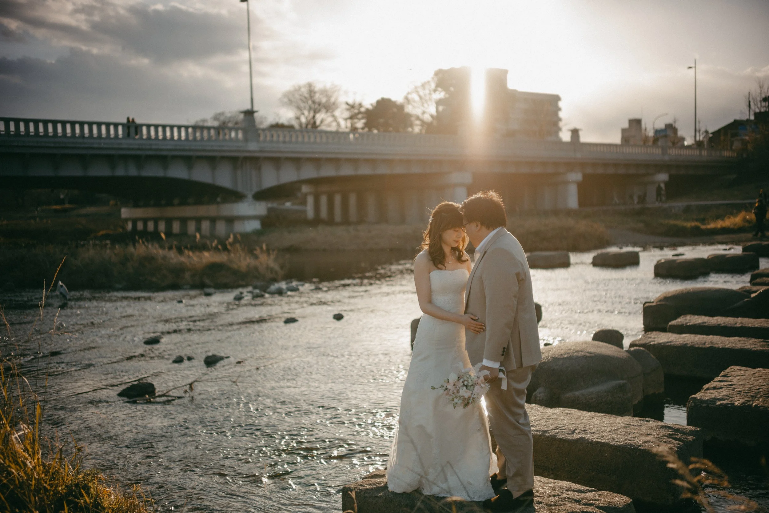 A bride and groom standing on rocks near a river during sunset amid a cityscape, holding hands and gazing at each other.