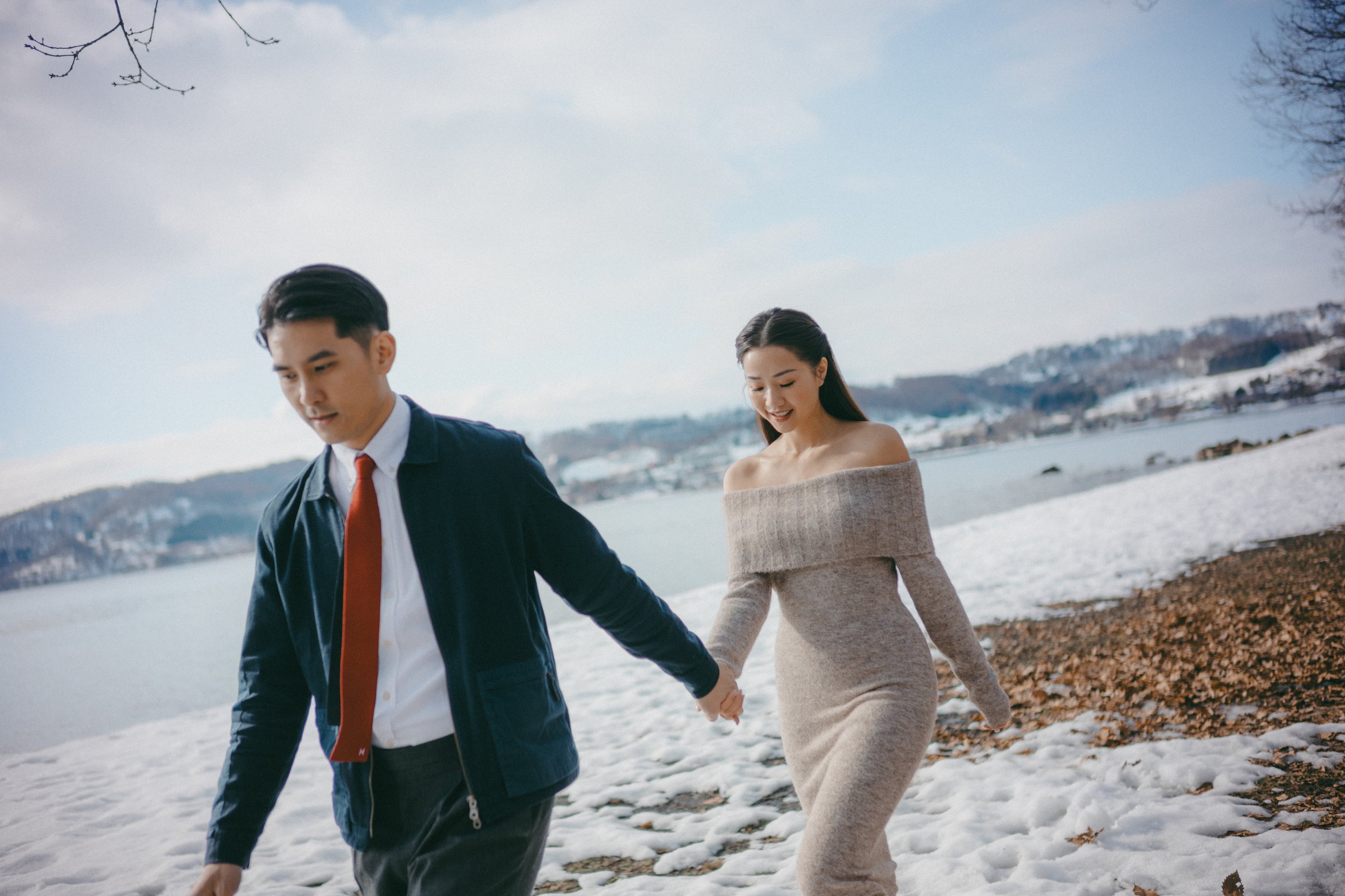 A young couple walking hand in hand along a snowy lakeshore on a clear day, with hills in the background. The man is wearing a navy jacket, white shirt, and red tie, while the woman is dressed in an off-the-shoulder beige dress.