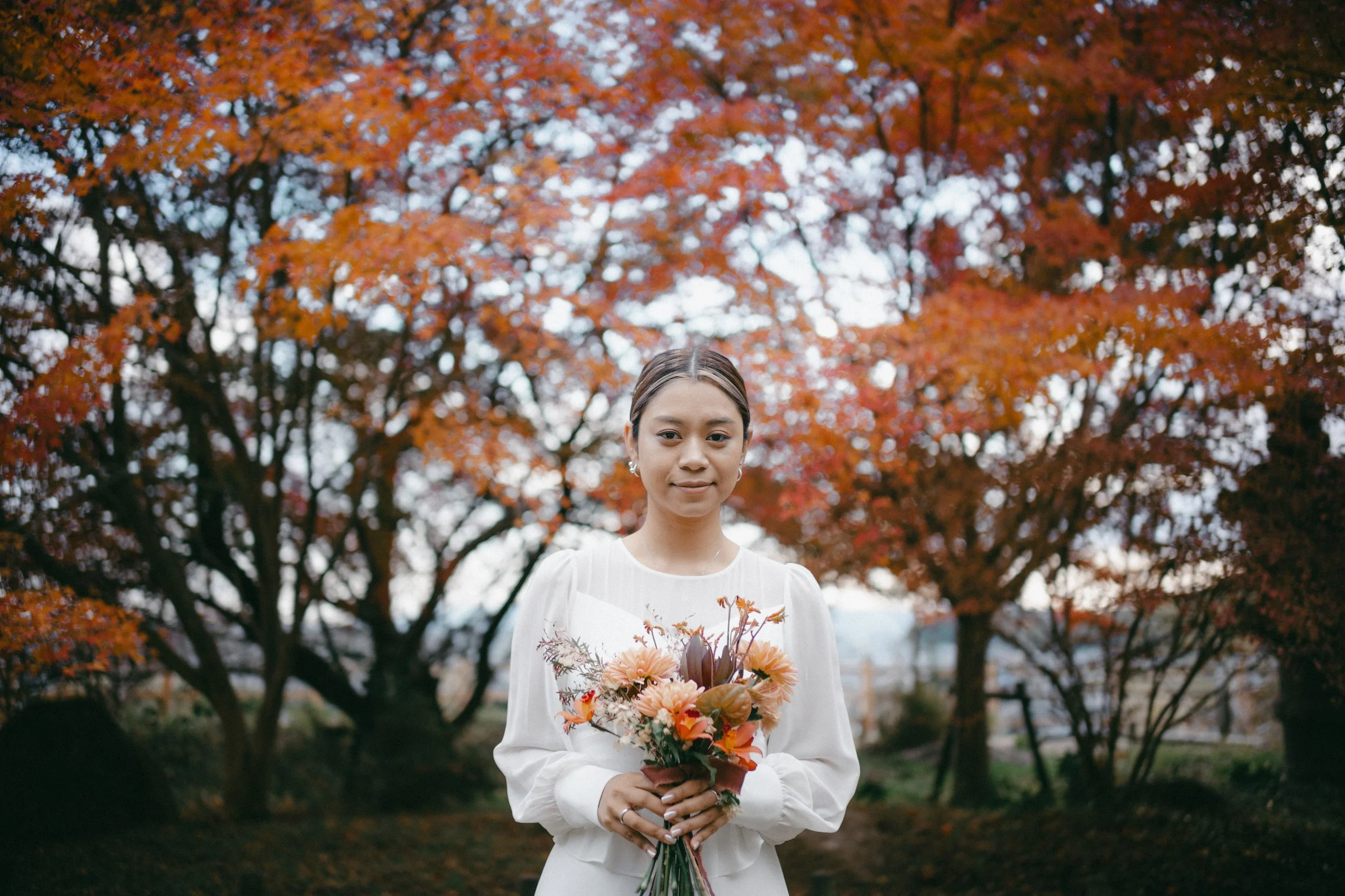 A woman in a white dress holding a bouquet of flowers in front of orange autumn trees.
