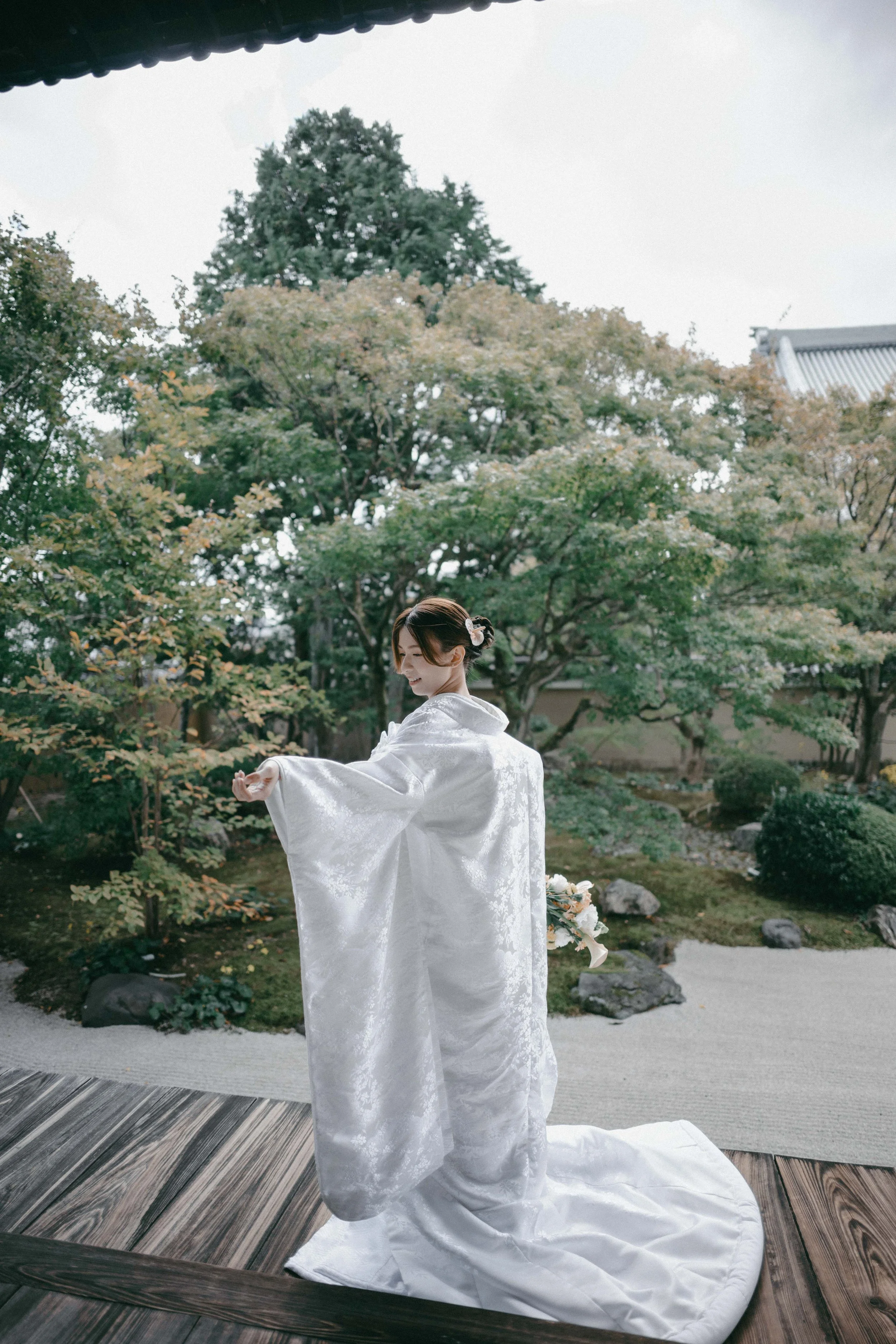 A woman dressed in a traditional white wedding kimono standing on a wooden deck in a Japanese garden.