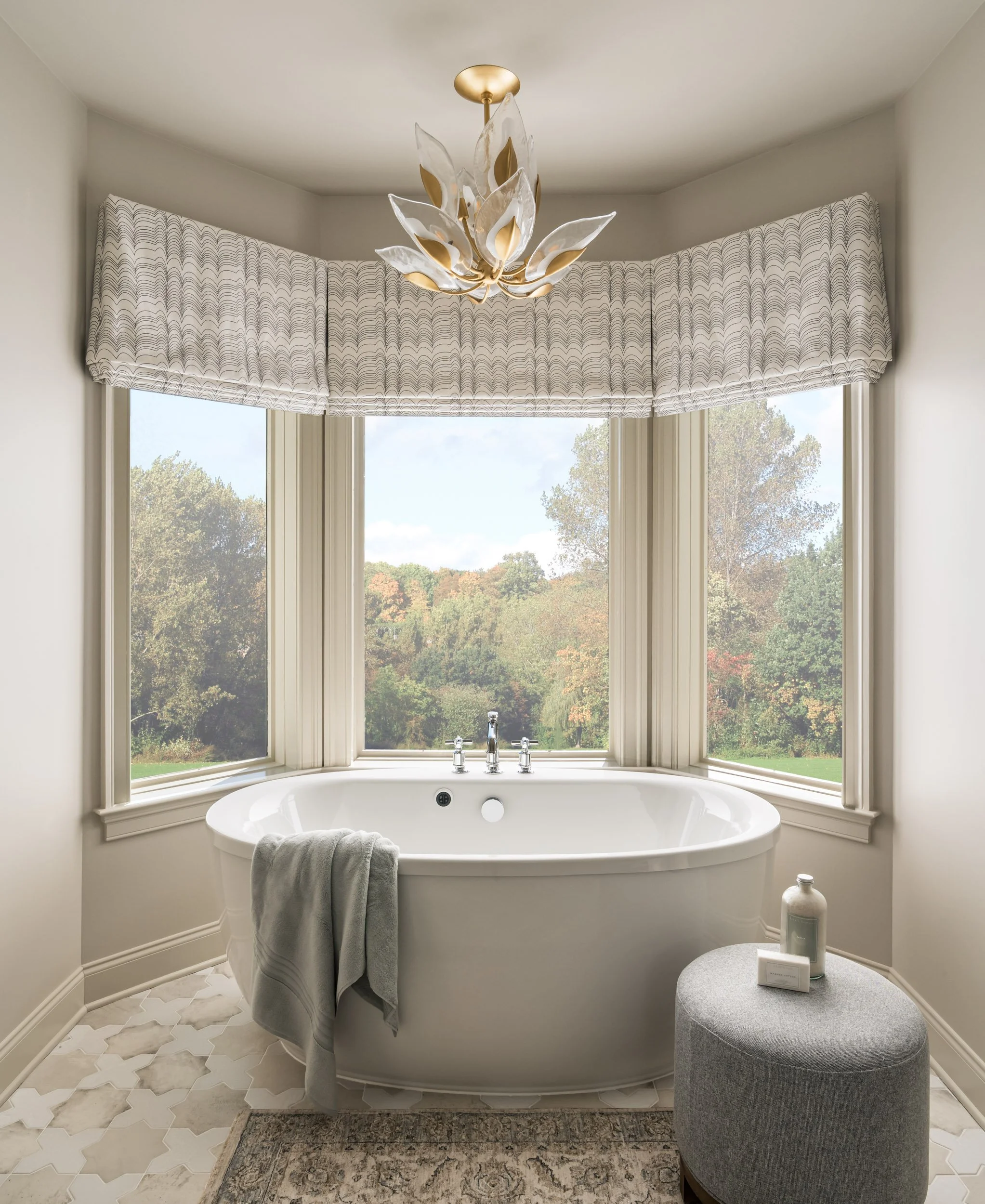 Luxurious bathroom with a white freestanding bathtub in front of large windows, a modern chandelier, patterned curtains, a fluffy towel, and a small ottoman with bath items.