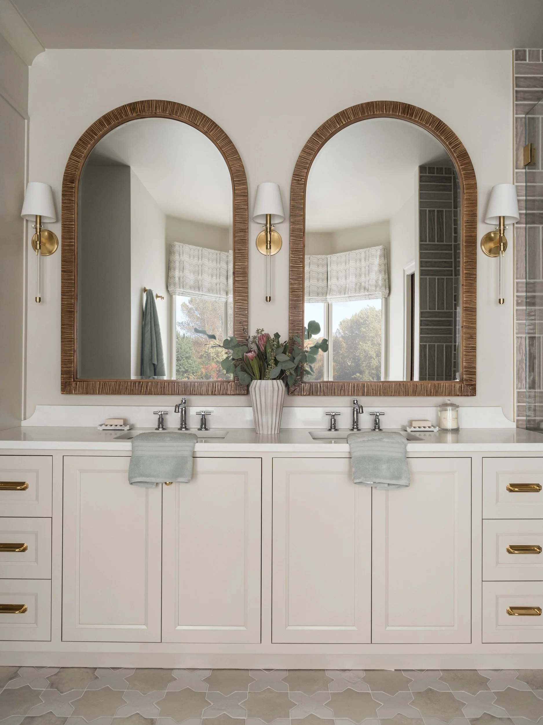 Elegant bathroom interior with a double sink vanity featuring two large arched mirrors, brass fixtures, and a flower vase. White cabinets with brass handles and soft green towels are visible. A window with sheer curtains is reflected in the mirrors.
