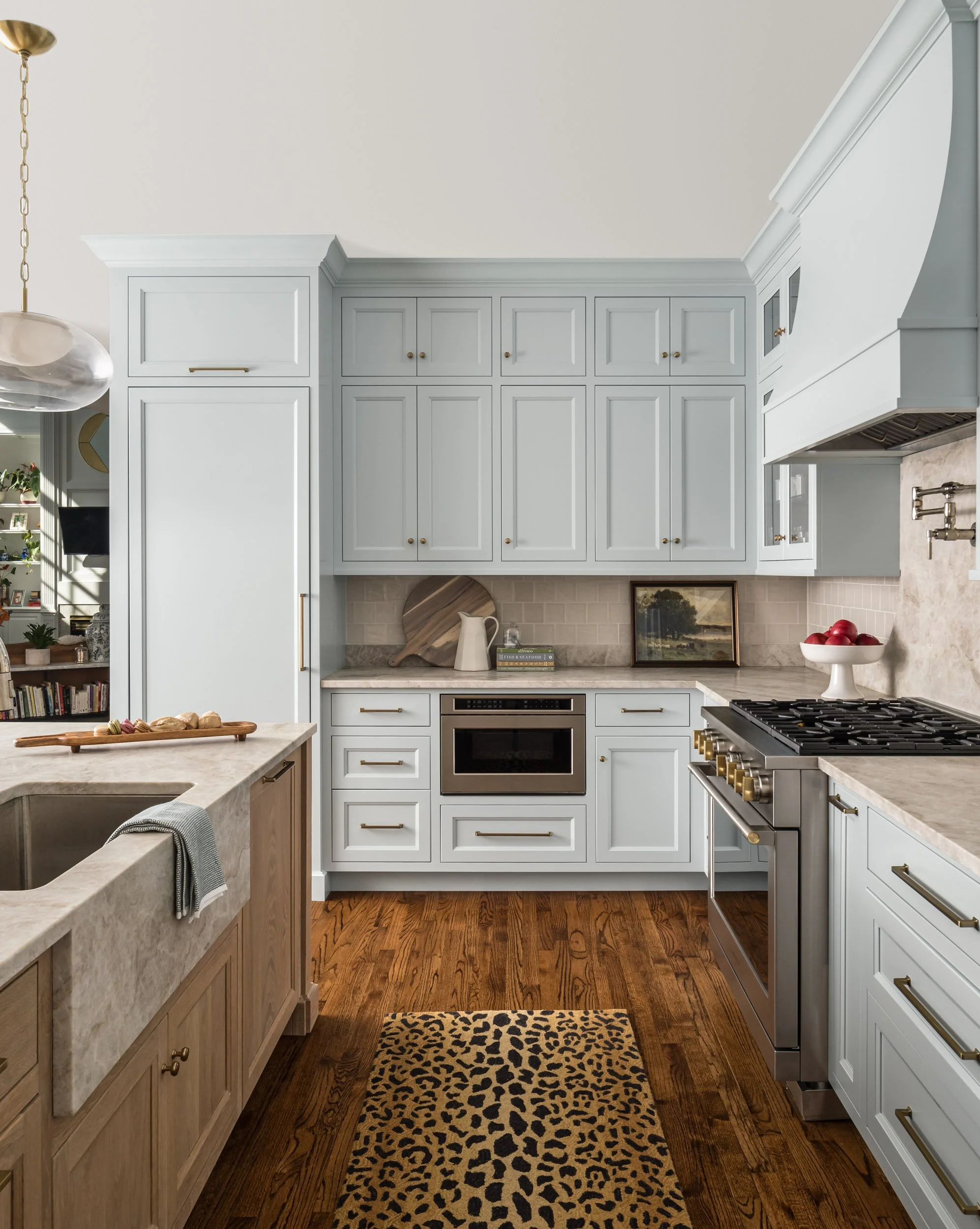 Modern kitchen with light blue cabinets, stainless steel appliances, marble countertops, a farmhouse sink, wooden floor, and a leopard print rug.