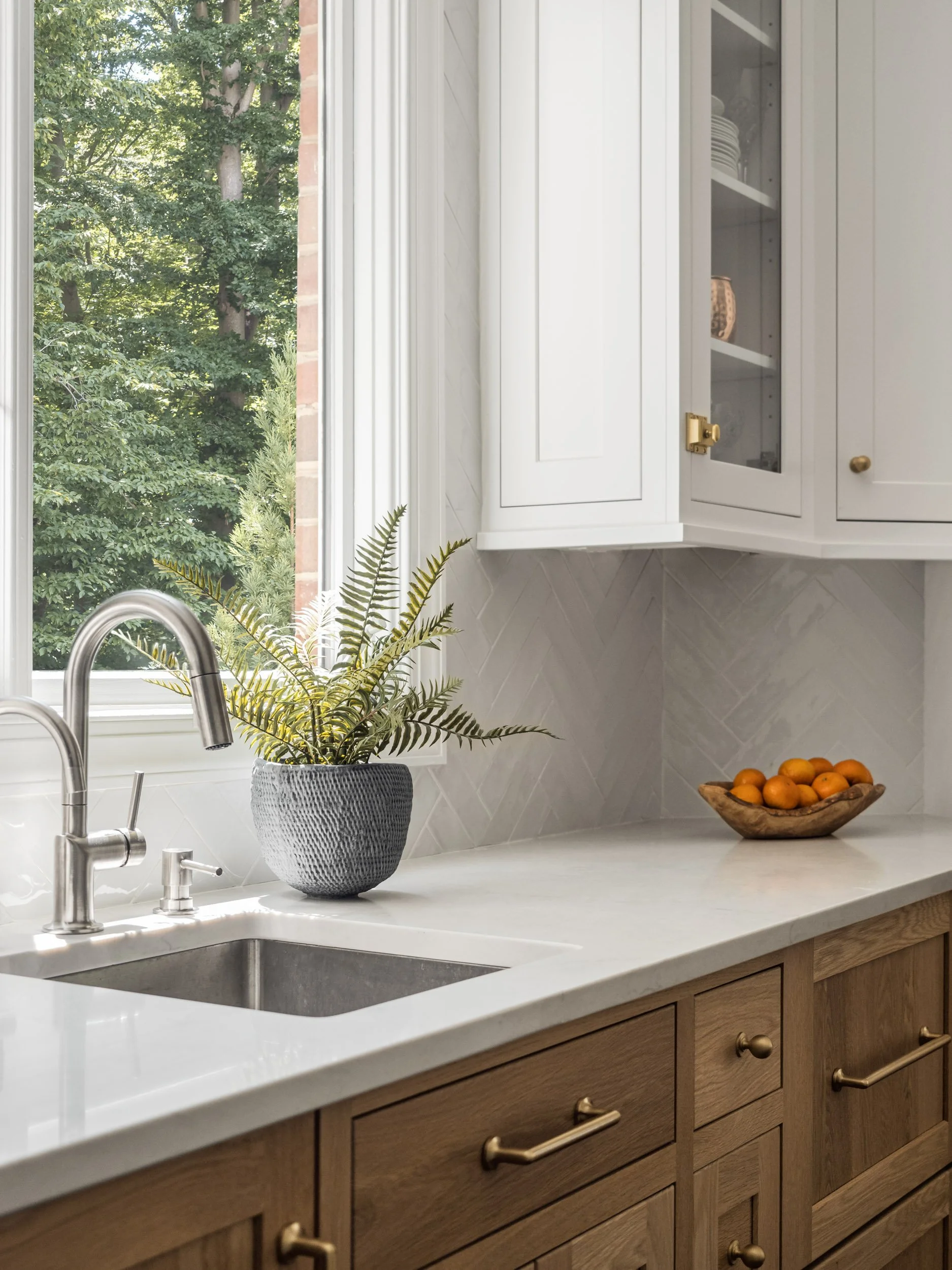 Modern kitchen with wooden cabinets, stainless steel sink, potted plant, bowl of oranges, and a large window with a view of trees.