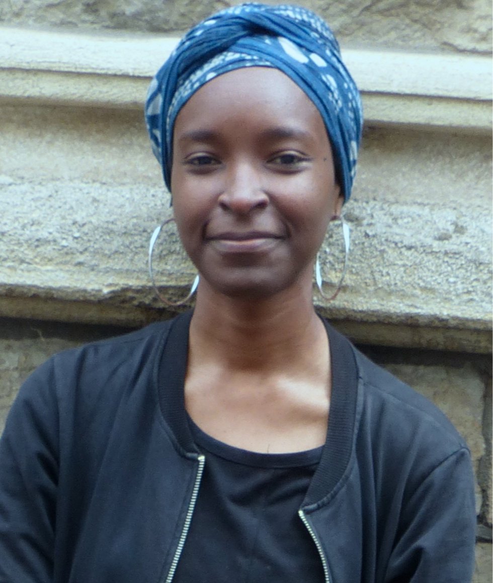 Photo of the artist, shoulder up. Artist wearing black clothing, silver hoop earrings and blue and white headwrap standing in front of a brick wall.