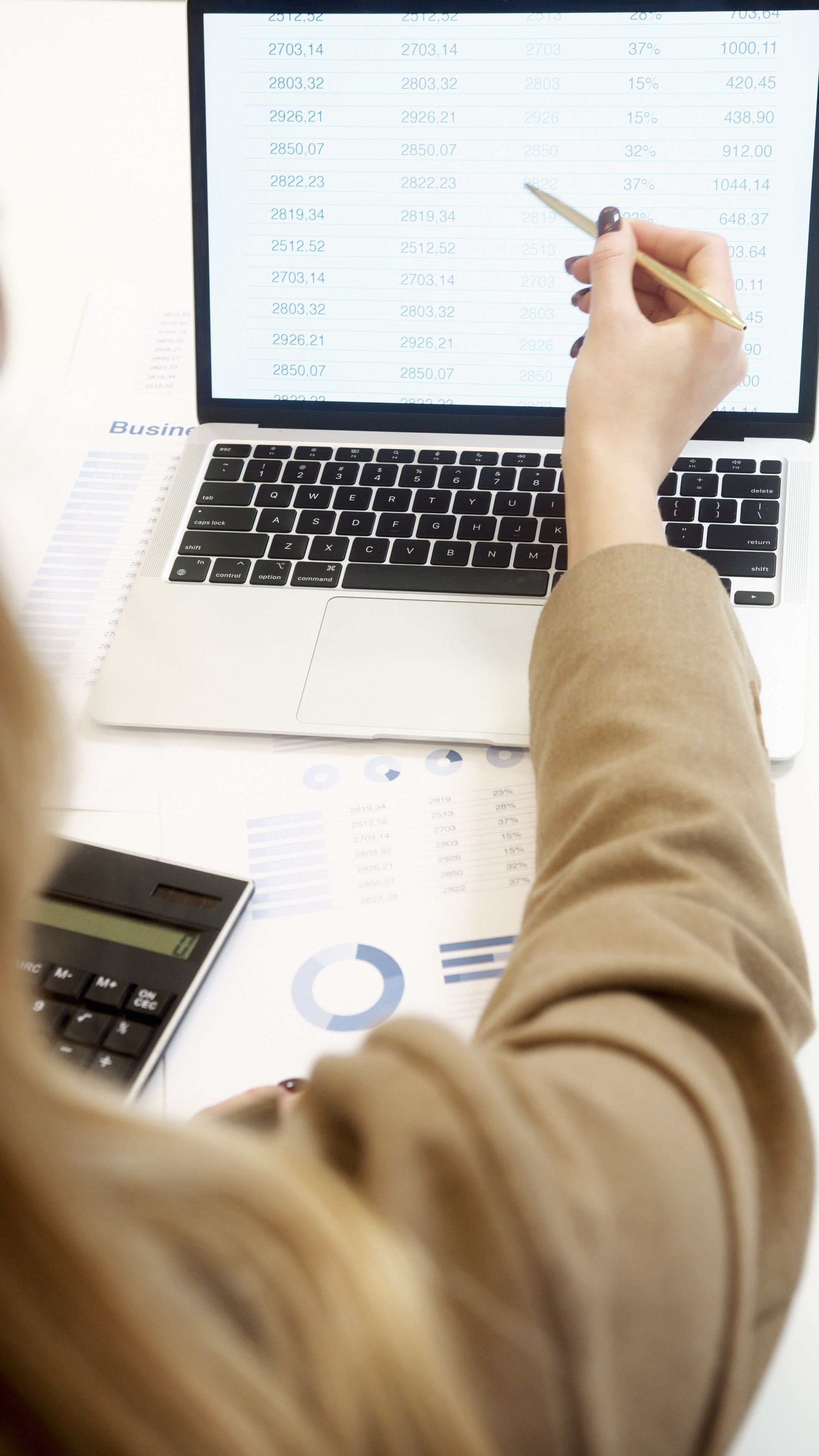 A person in a brown sweater is working on a spreadsheet on a laptop, with financial charts and a calculator on the desk.