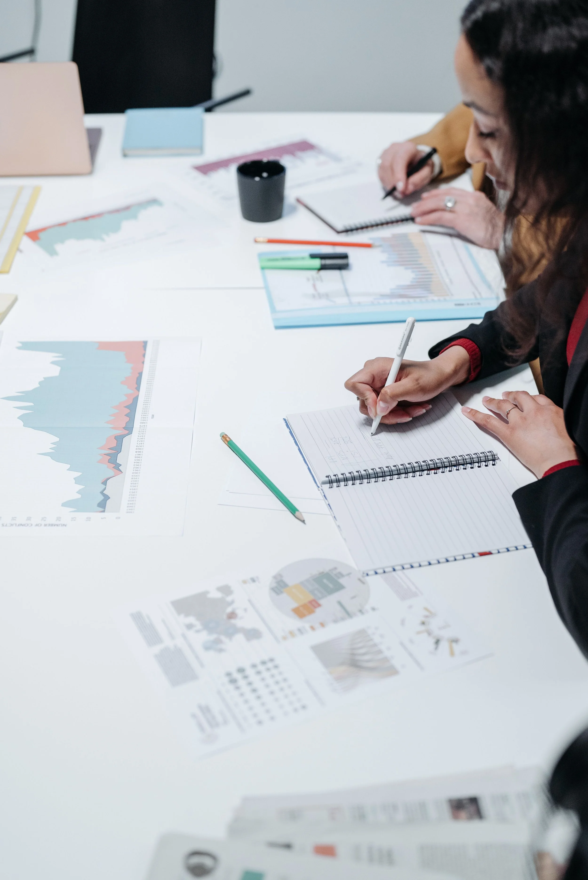 People sit at a white table covered with various printed graphs, charts, and documents. One person writes in a notebook, while others work with pens and highlighters. The scene appears to be in a meeting or collaborative work session.