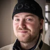 Close-up of a man with light skin, blue eyes, and a beard, wearing a black beanie and a beige fleece jacket, standing indoors with a slight smile.