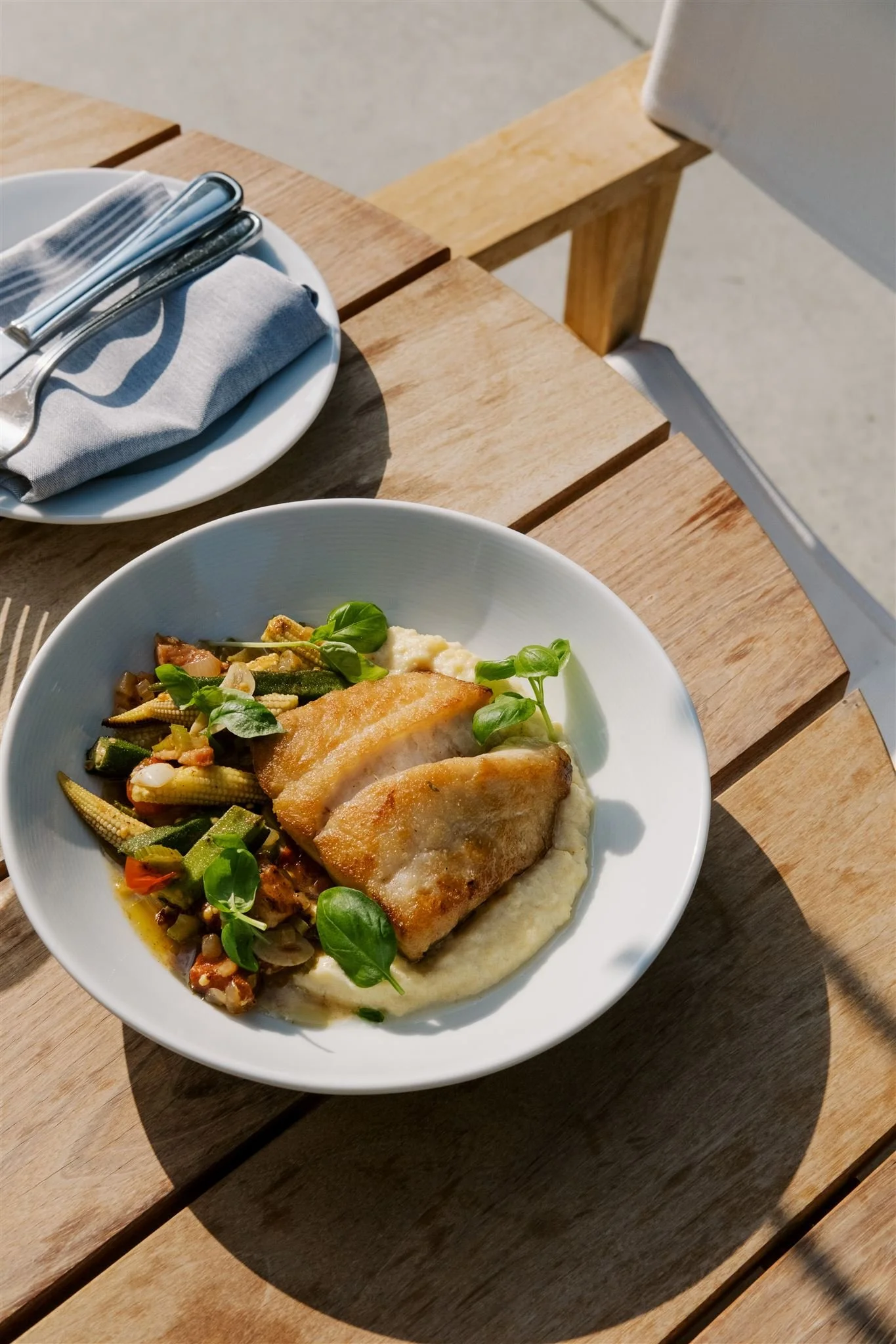 A white bowl with two pieces of fish on a bed of mashed potatoes, served with sautéed mixed vegetables and garnished with fresh herbs, placed on a wooden table outdoors.