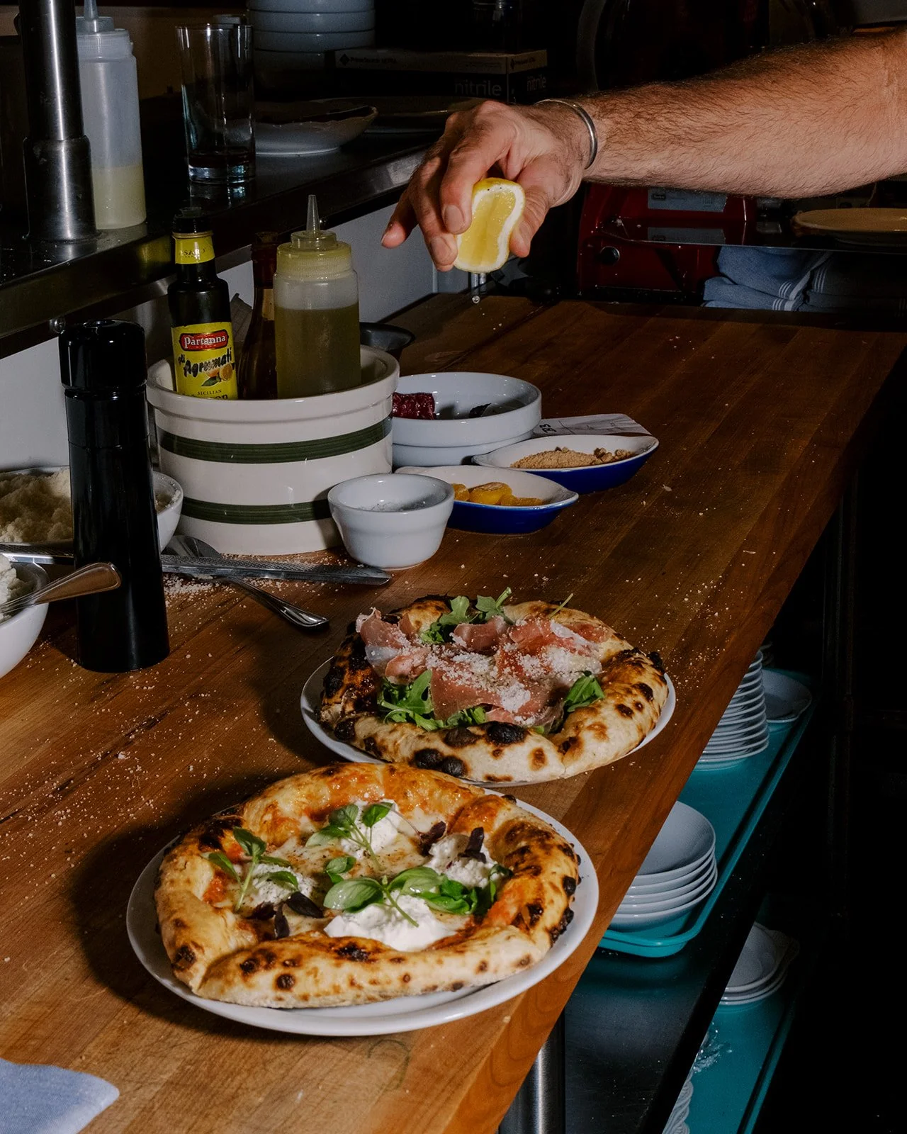 A person squeezing lemon juice onto food at a restaurant counter with pizzas and various condiments visible.