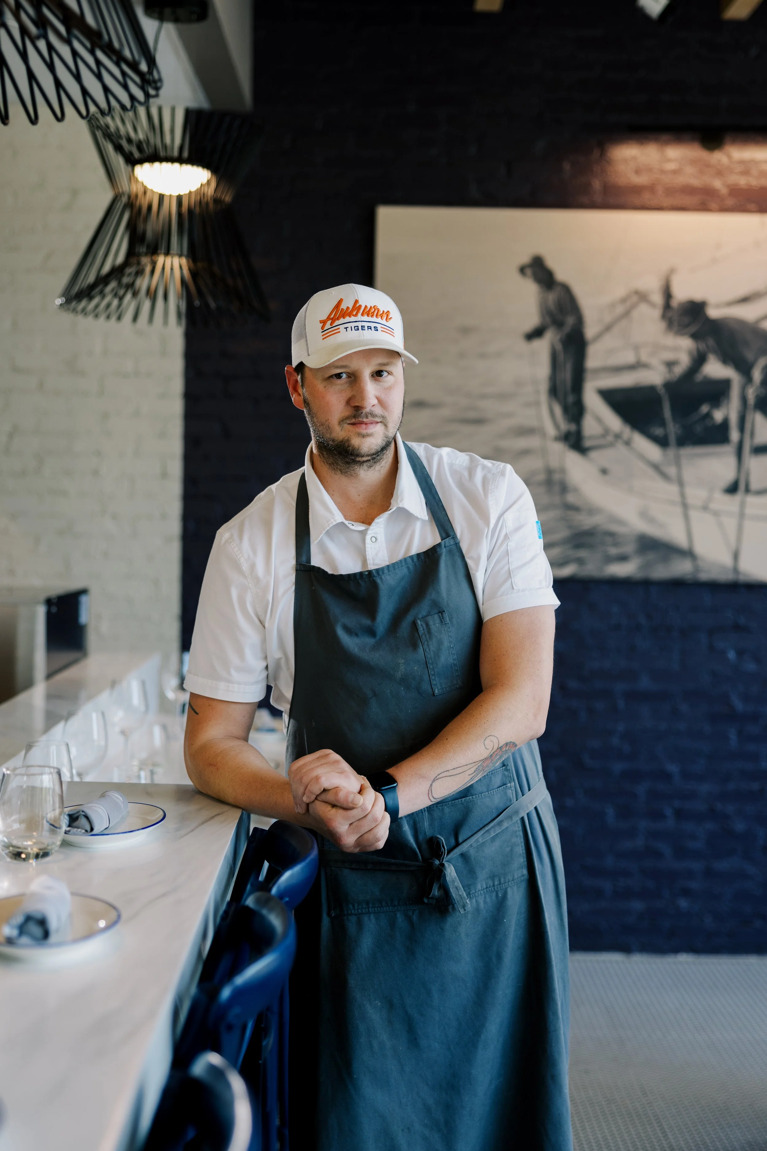 A man wearing a white cap with 'Auburn Tigers' logo, white shirt, and black apron stands next to a dining counter with glasses and napkins in a modern restaurant.
