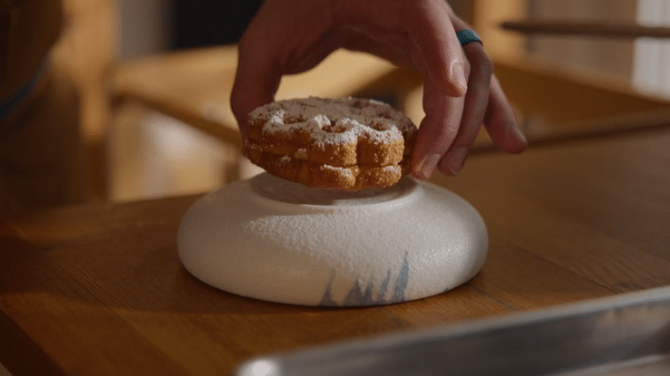 A hand placing a crumbly cookie on a white, textured ceramic plate on a wooden table.