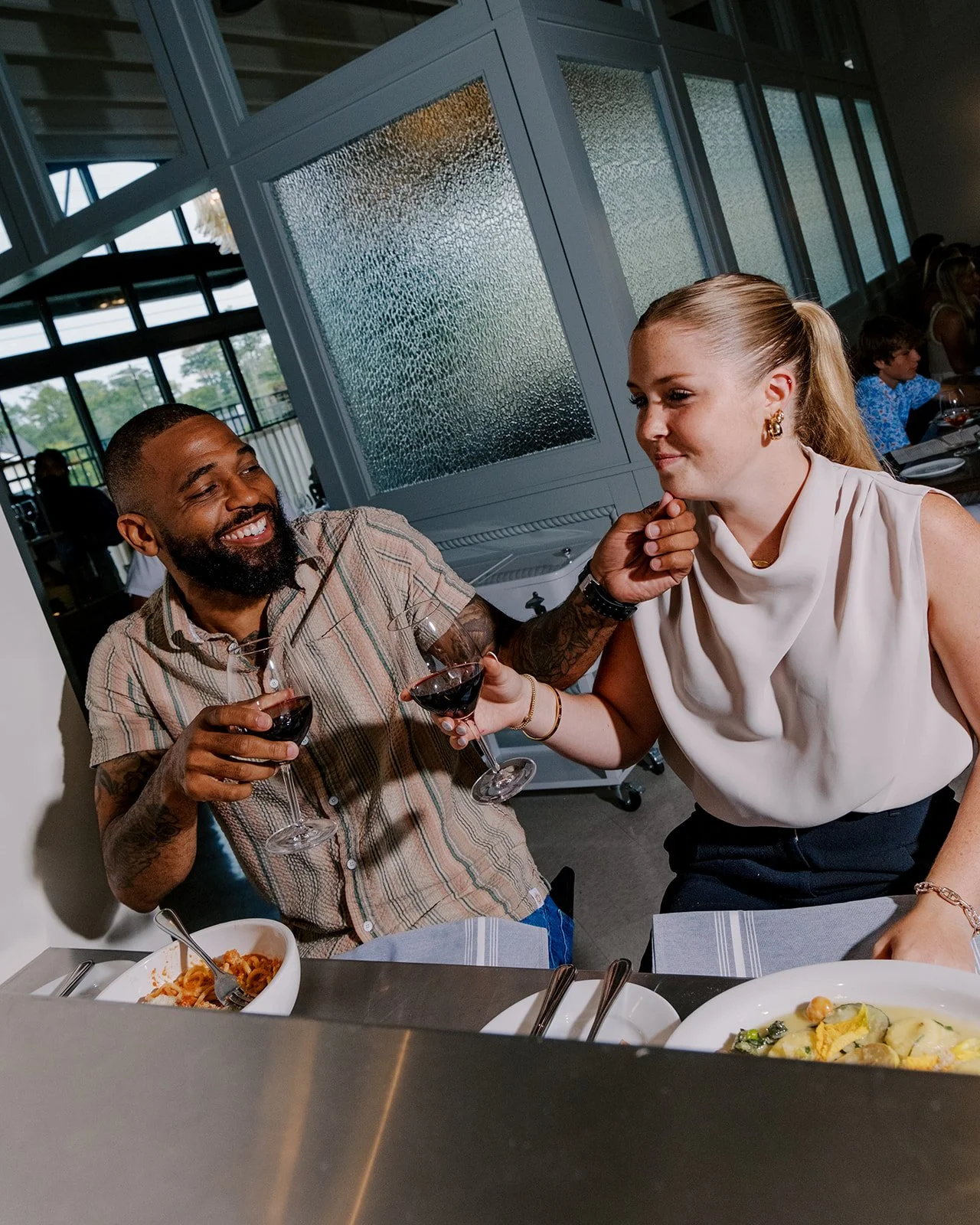 A man and woman at a restaurant, smiling and toasting with glasses of red wine, with plates of pasta on the table.