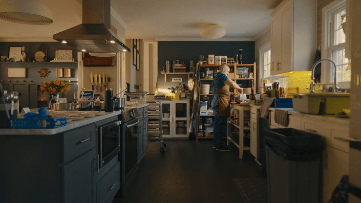 A person in an apron working in a well-lit kitchen with white cabinets, dark blue walls, and a window. The kitchen has various cooking utensils, containers, and open shelving.