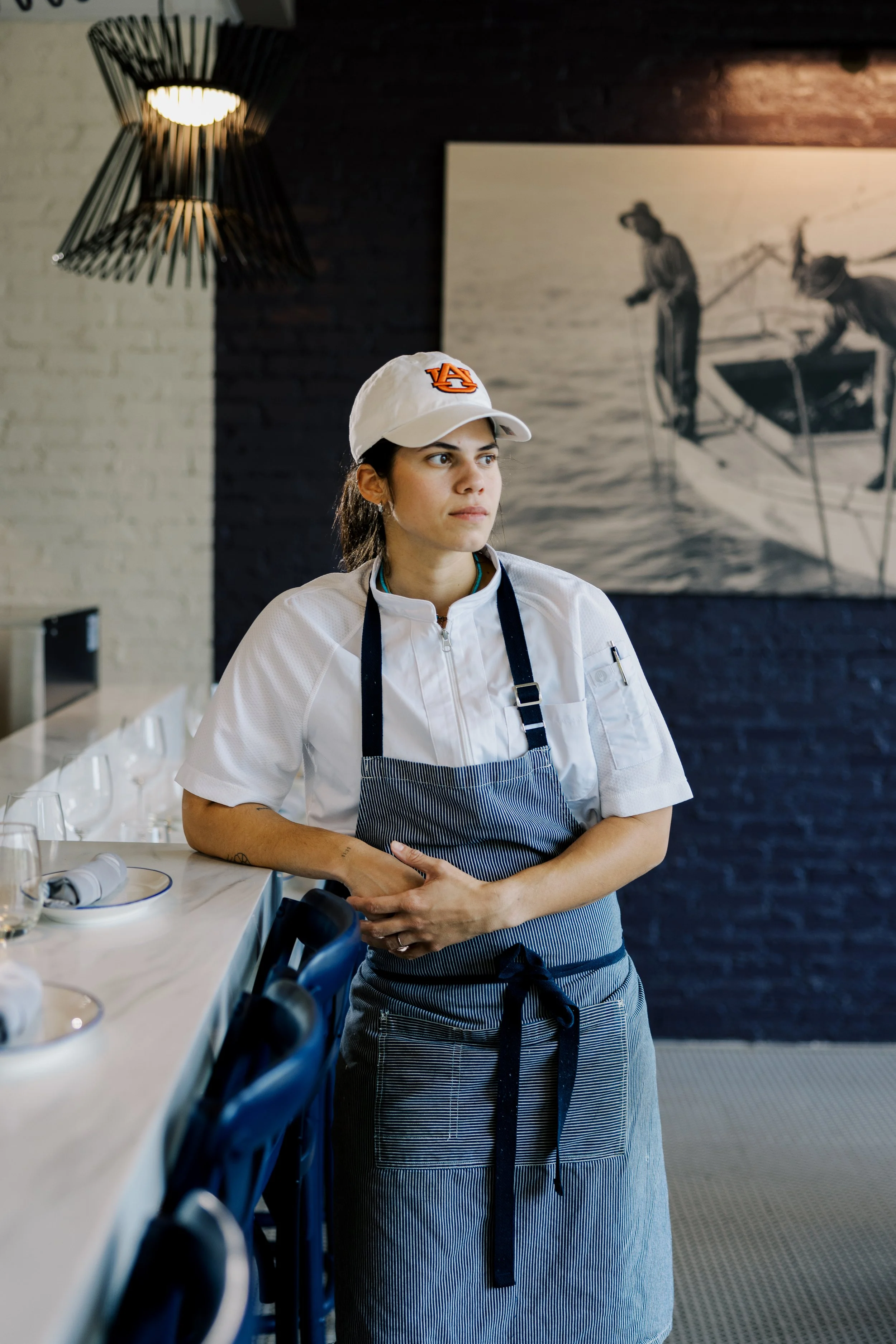 A female chef in a white Auburn University cap and striped apron stands by a restaurant counter, looking to the side with her hands clasped in front of her. There is a drawing of workers on a boat in the background.