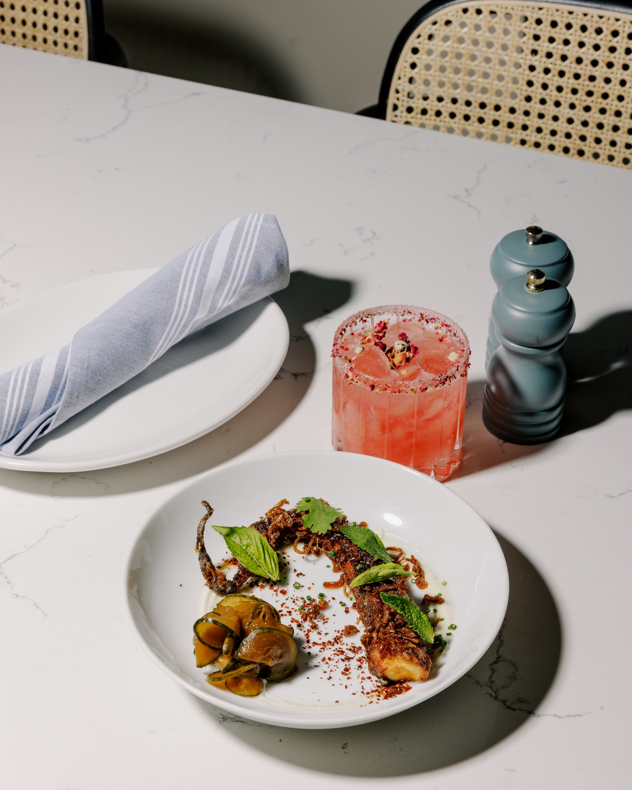 A white marble table set with a white plate holding a grilled meat skewer garnished with herbs and spices, and a side of pickled vegetables. There is a pink cocktail in a glass with salt on the rim, and salt and pepper shakers. A folded napkin rests 
