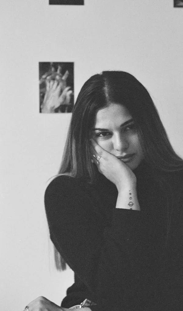 Black and white photo of a woman with long dark hair posing with her hand resting on her face, in front of a wall with a small photo of hands playing a musical instrument.