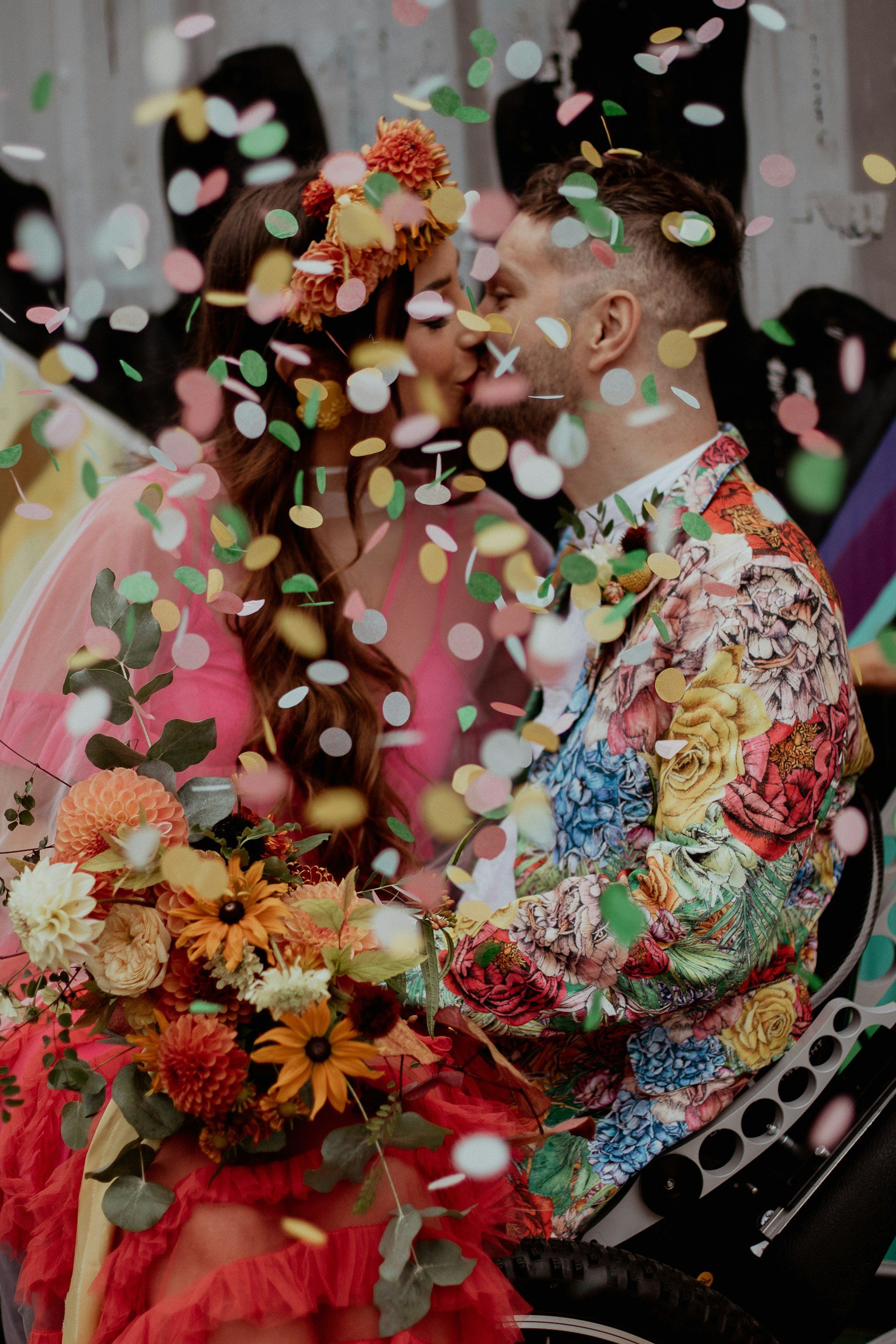 A couple is kissing at their wedding, surrounded by falling confetti, with a bride holding a bouquet of flowers and wearing a floral crown, and both dressed in vibrant, floral-themed attire.