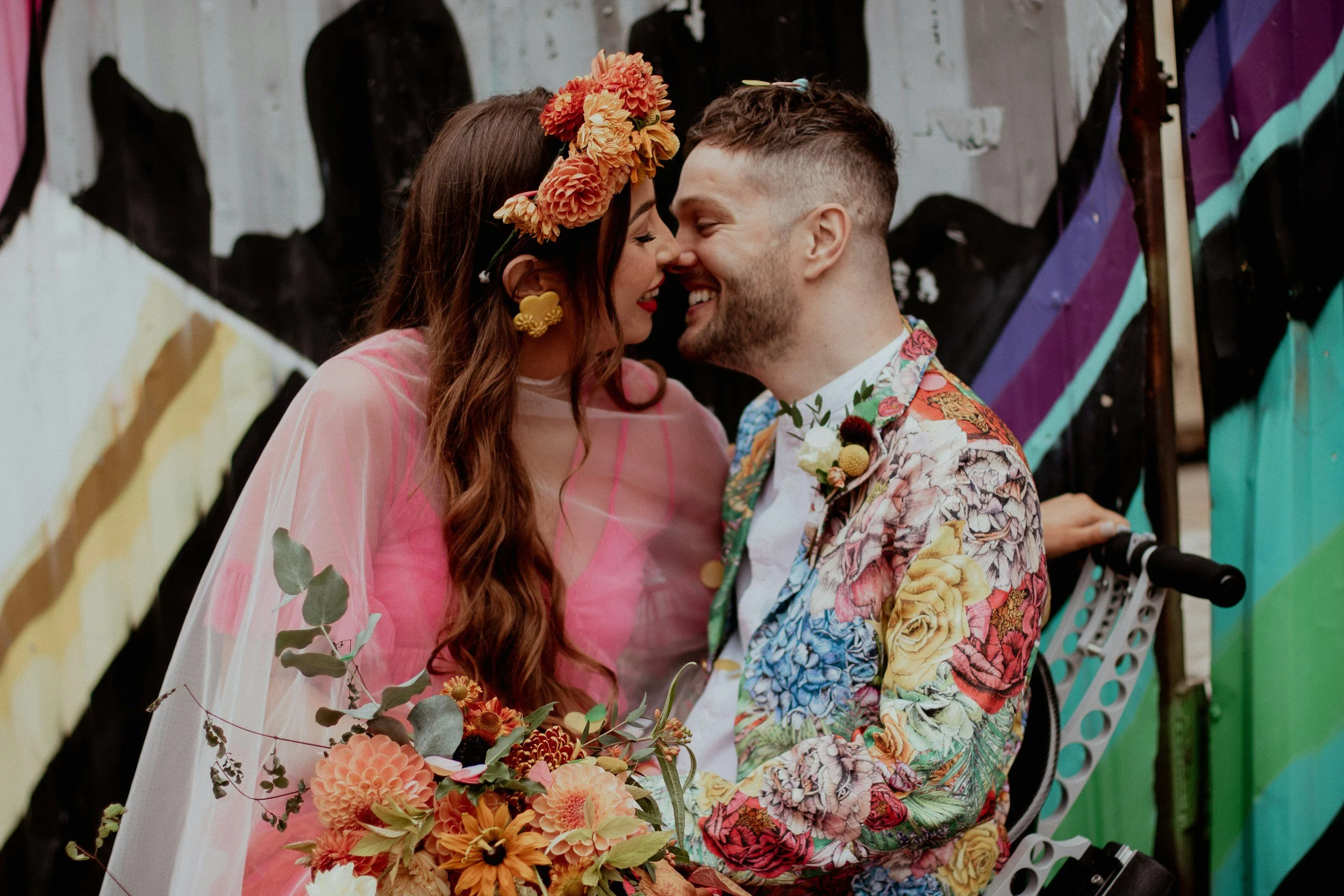 A couple smiling and touching noses at their wedding. The bride has long, wavy hair and wears a pink dress with a floral headpiece and earrings. The groom has short hair, a beard, and wears a colorful floral suit jacket. They hold a bouquet of flowers, and there is a colorful painted wall in the background.