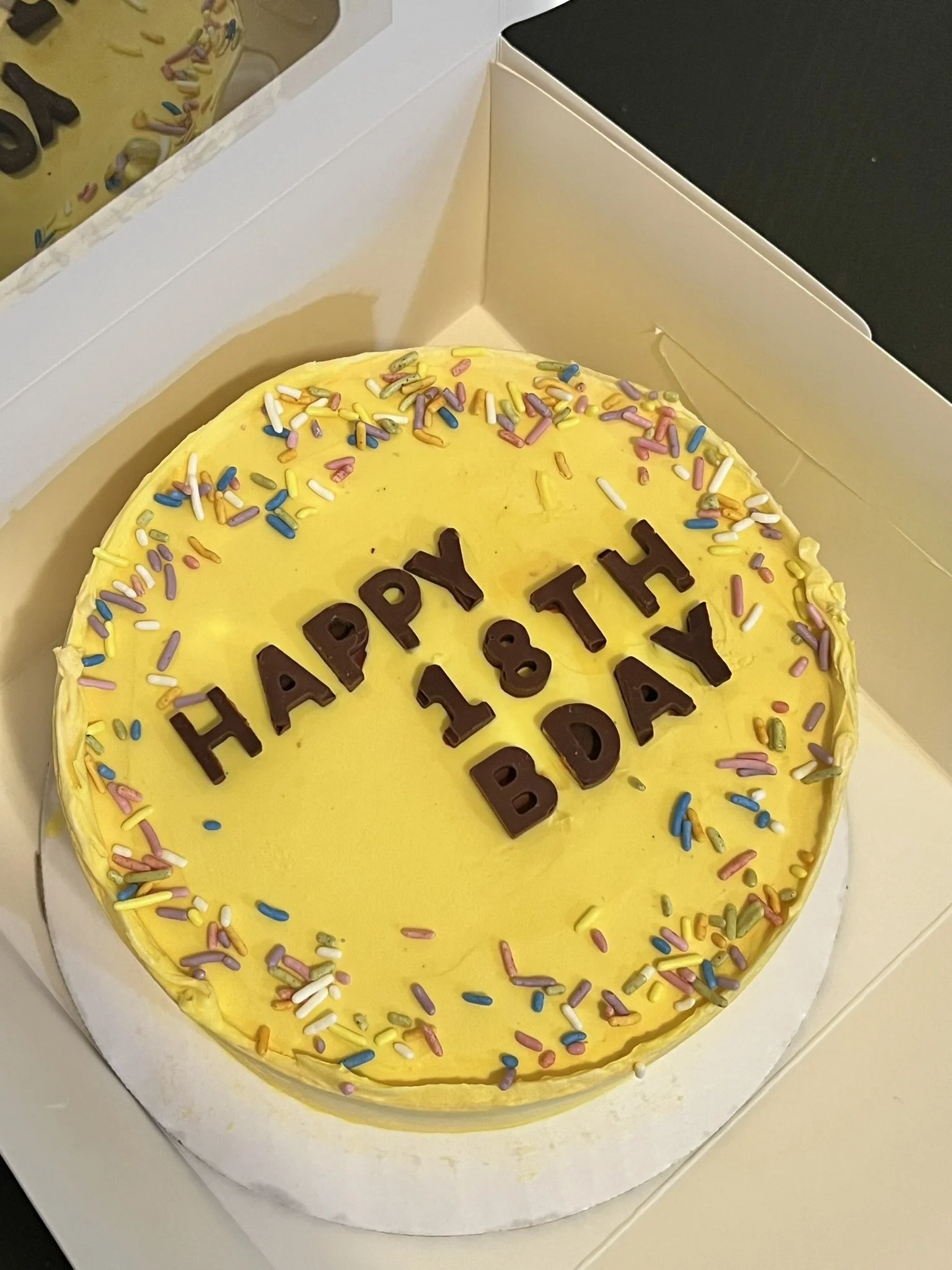 Yellow birthday cake with colorful sprinkles and chocolate letters spelling 'HAPPY 18TH BIRTHDAY' in a white box