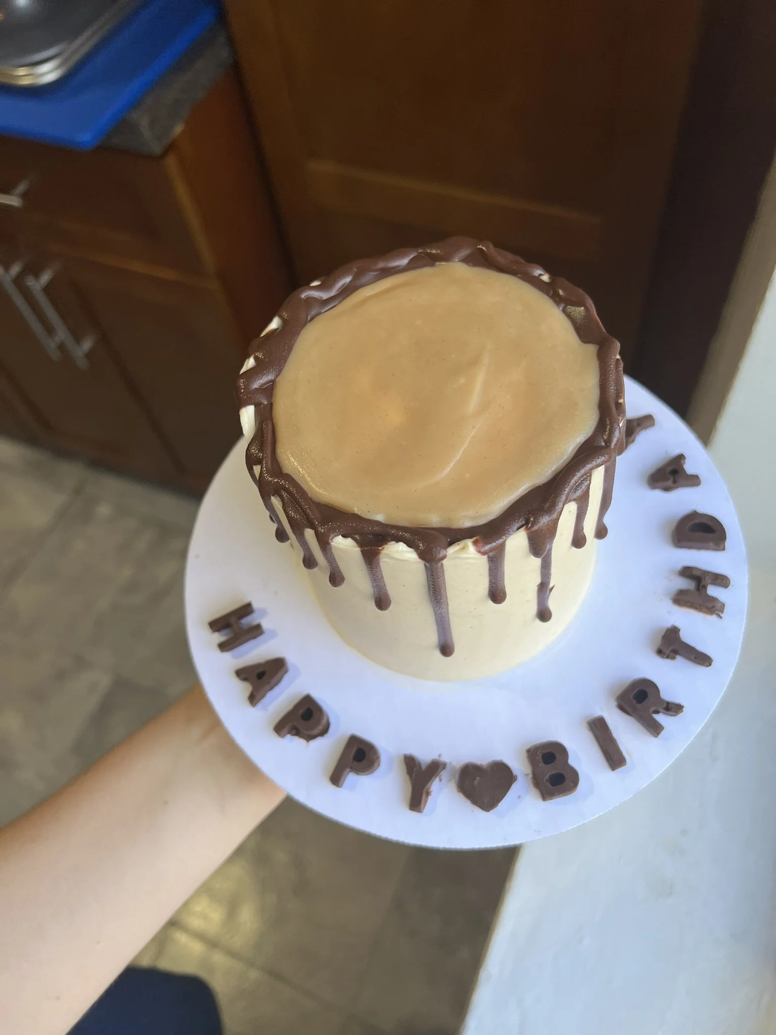 A birthday cake with caramel topping on a white circular board with a chocolate border, surrounded by chocolate letters spelling 'HAPPY BIRTHDAY'.